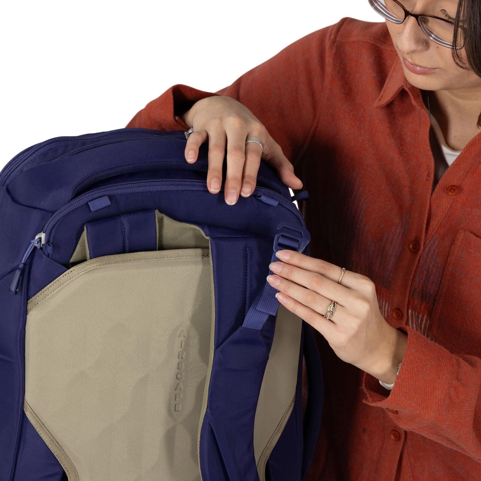 A person wearing a rust-colored shirt is adjusting a blue and tan backpack. Their hands are shown tightening a strap near the top of the pack. The back panel is exposed, featuring padded sections and the word "AIRSCAPE" molded into the tan material. Zippe