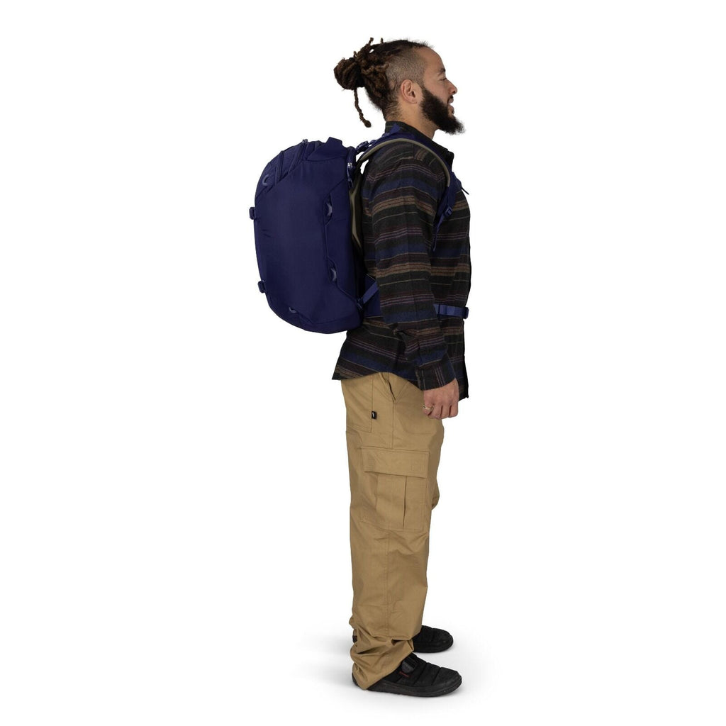 A person is shown in side profile wearing a dark blue backpack with multiple attachment points and straps. They are dressed in a dark striped long_sleeve shirt, tan cargo pants, and black shoes. The backpack sits snugly with chest and waist straps visible