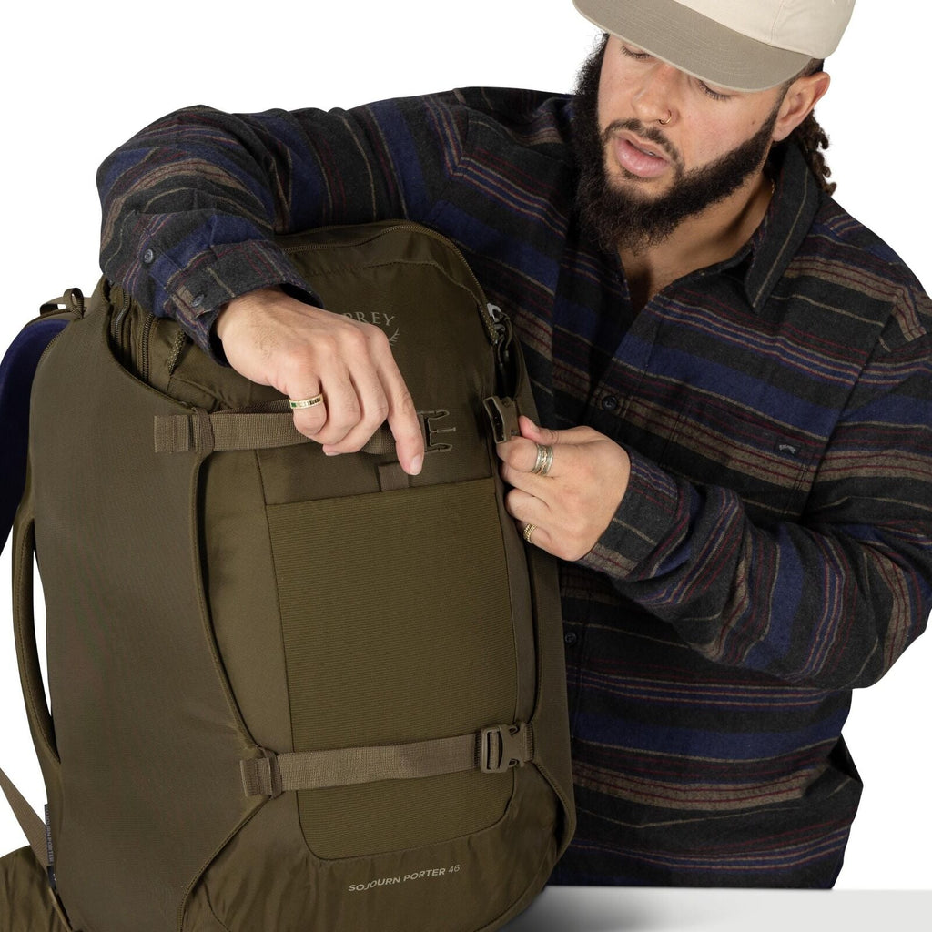 A person wearing a striped flannel shirt adjusts a buckle on an olive-green travel backpack. The pack has compression straps on the front and sides, a flat front panel pocket, and rounded edges. The logo "Osprey" is faintly visible near the top, and "Sojo