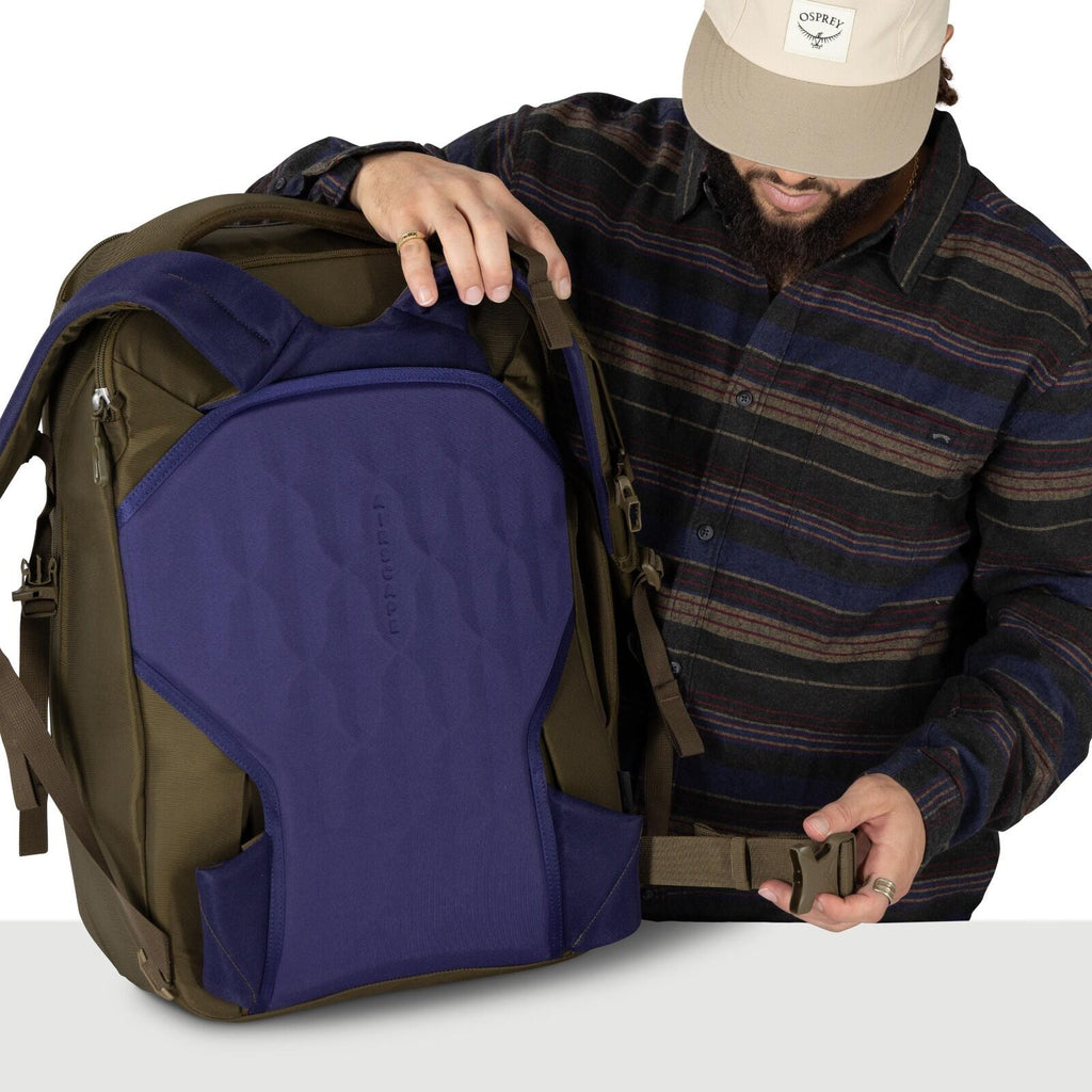 A person wearing a striped flannel shirt and a beige cap is holding a large backpack. The pack is olive green with a prominent purple back panel that says "AirScape." The person is adjusting or fastening the hip belt buckle while holding one shoulder stra