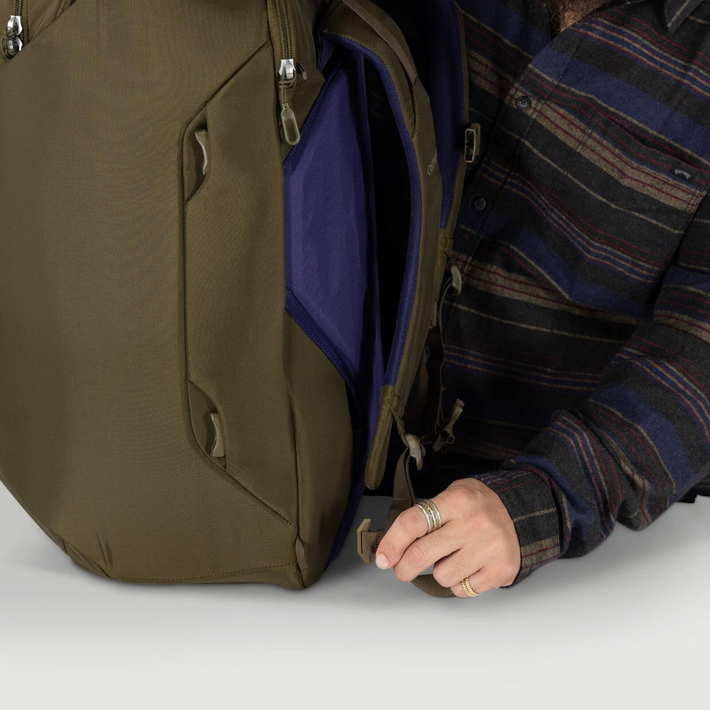 A person adjusts a strap on an olive-green backpack that is partially unzipped. The interior shows a purple compartment panel and tan zipper pulls. The person wears a dark, striped flannel shirt and multiple rings. The scene focuses on the bag's side prof