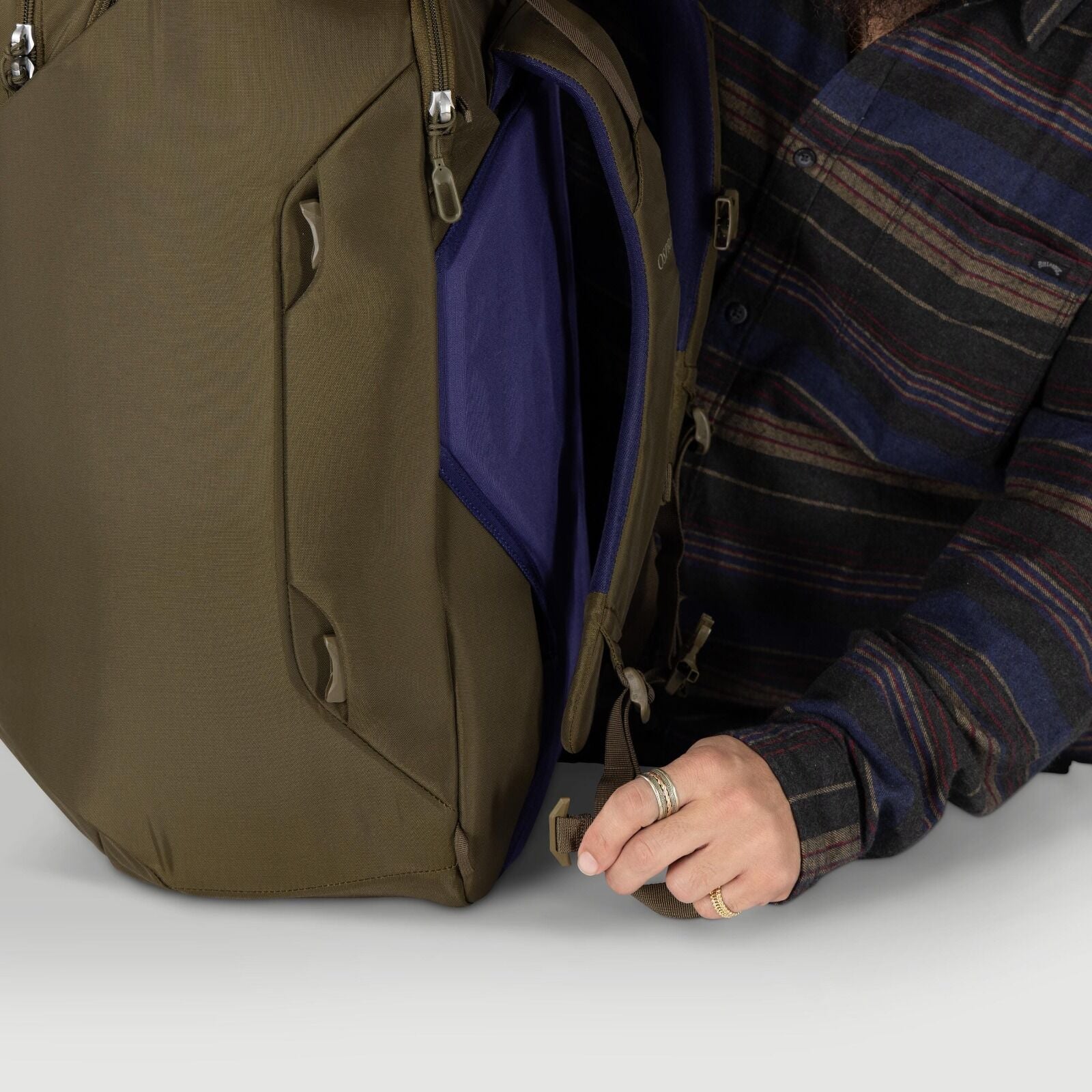 A person adjusts a strap on an olive-green backpack that is partially unzipped. The interior shows a purple compartment panel and tan zipper pulls. The person wears a dark, striped flannel shirt and multiple rings. The scene focuses on the bag's side prof