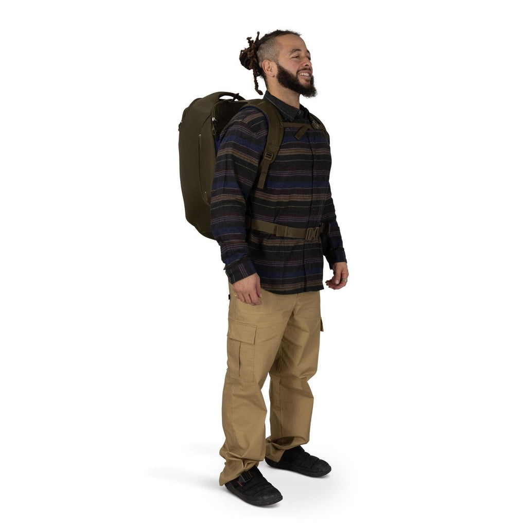 A person stands in a three-quarter pose wearing a large olive-green backpack with padded straps and a waist belt. They're dressed in a dark striped button-up shirt, tan cargo pants, and black sneakers. The backpack looks structured and travel-oriented, ex