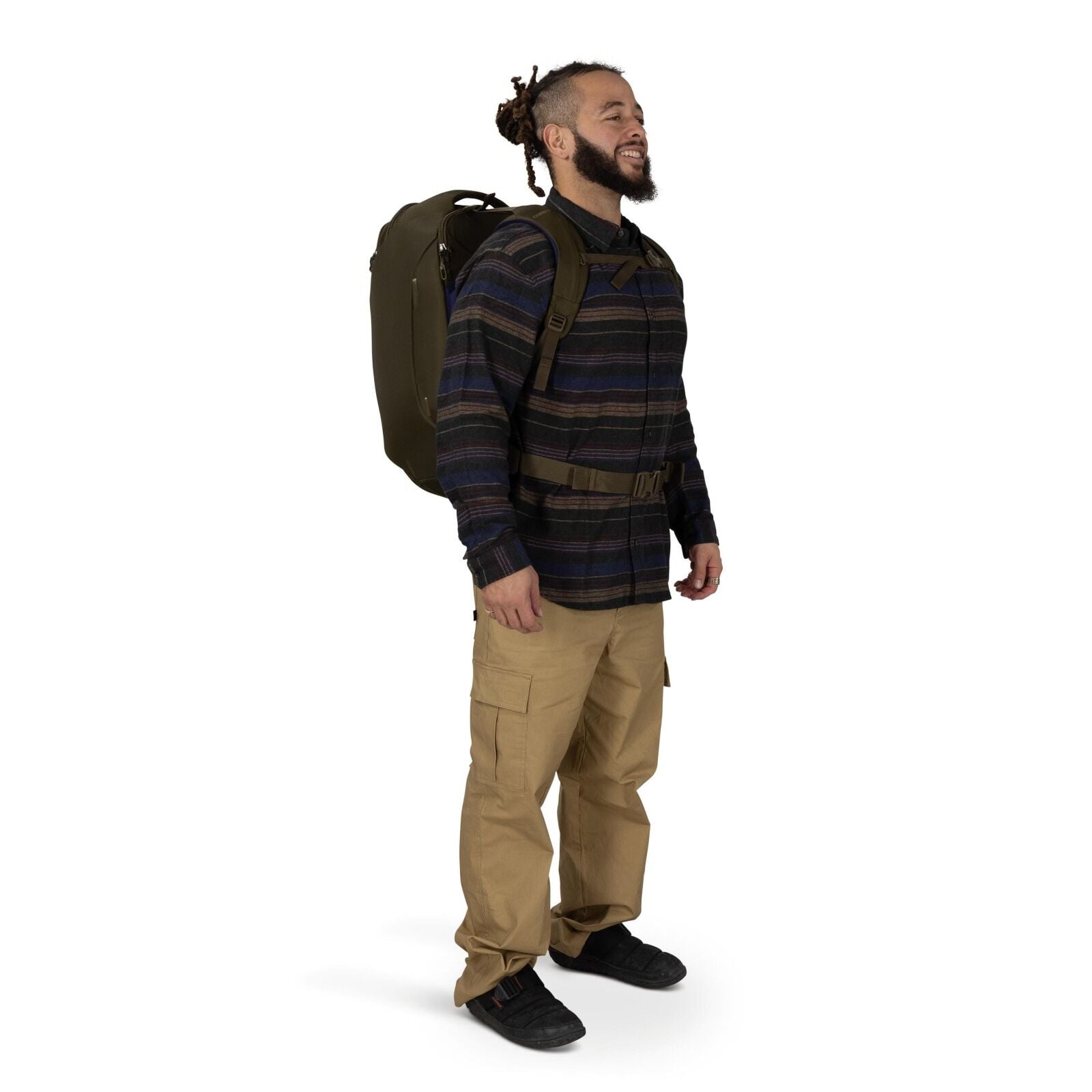 A person stands in a three-quarter pose wearing a large olive-green backpack with padded straps and a waist belt. They're dressed in a dark striped button-up shirt, tan cargo pants, and black sneakers. The backpack looks structured and travel-oriented, ex