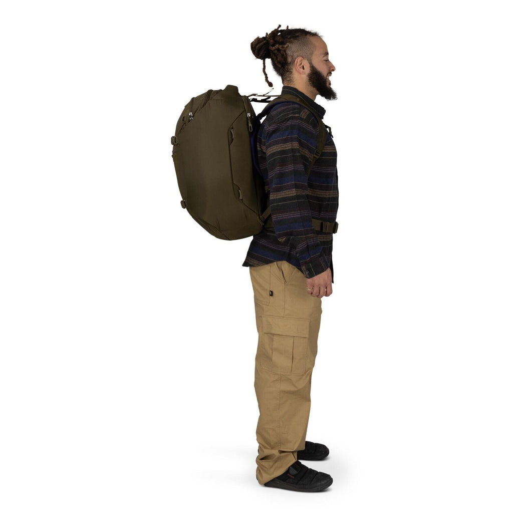 A person is shown in a side profile wearing an olive-green travel backpack. They are dressed in a dark striped button-up shirt, tan cargo pants, and black shoes. The backpack appears large and structured, with side compression straps and a hip belt for su