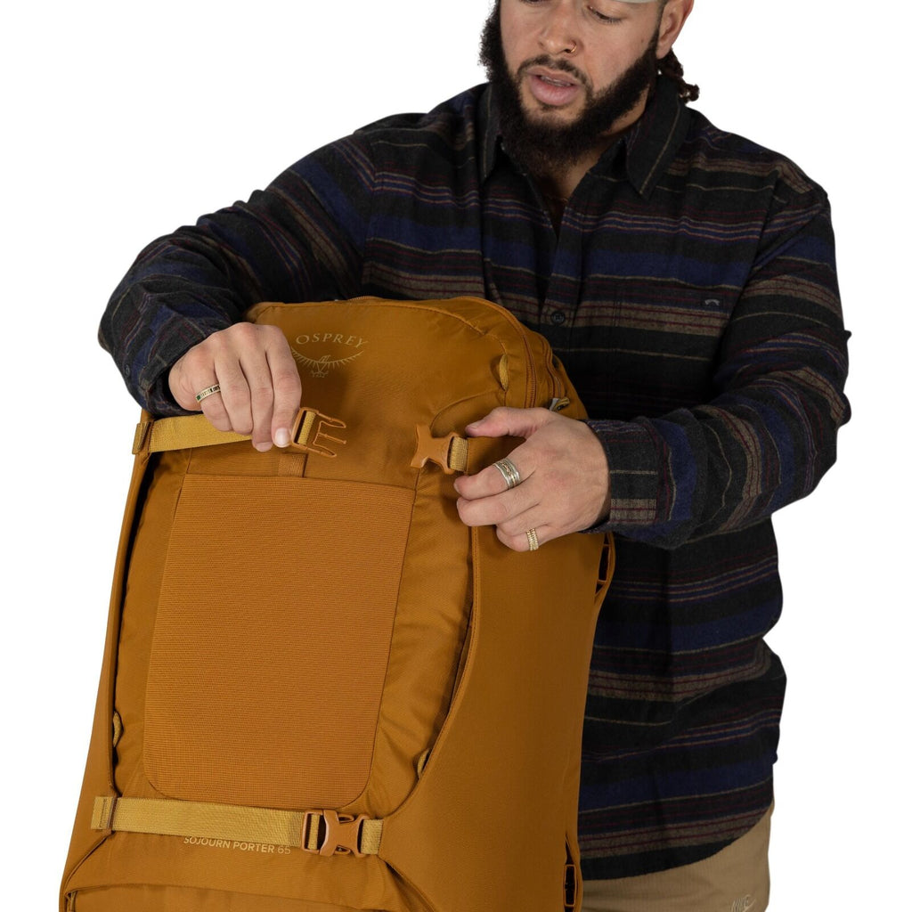 A person wearing a dark, striped button-up shirt is adjusting the side compression strap on a large, mustard-colored Osprey backpack. The pack features a front panel with a textured rectangular area and visible straps and buckles, with "Sojourn Porter 65"