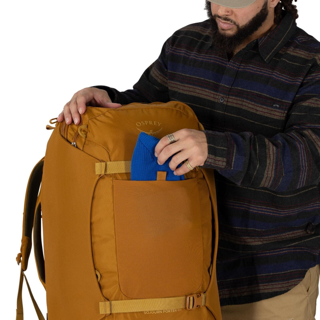 A person wearing a striped button-up shirt is placing a blue item into the front shove-it pocket of a large ochre-colored Osprey backpack. The pack has compression straps and a top zipper, with the Osprey logo visible near the top. The scene focuses on th