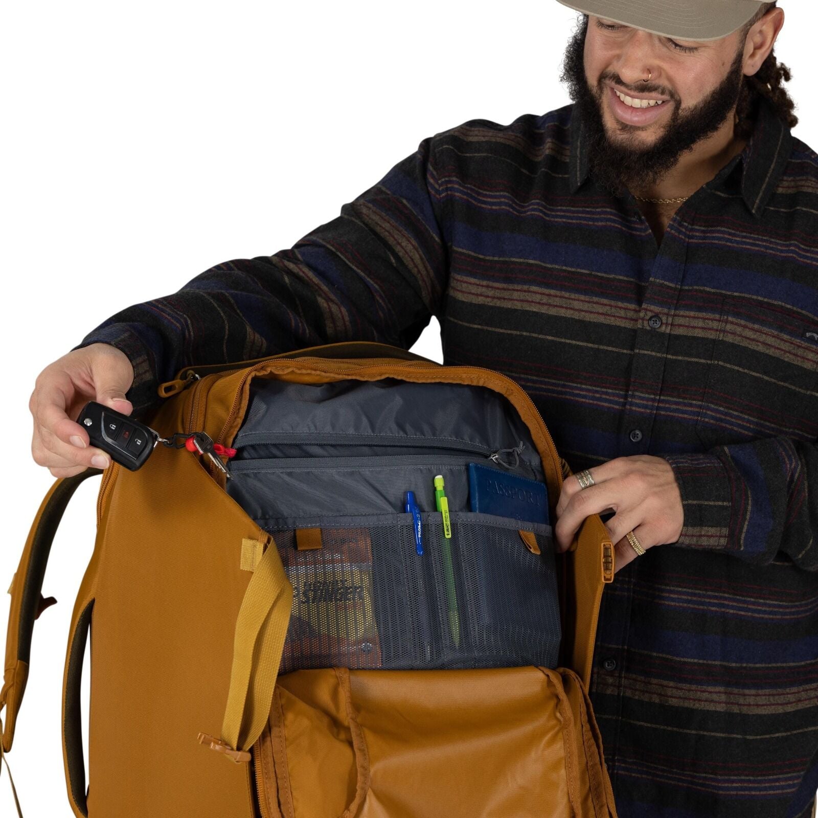 A person opens a mustard-colored backpack to show its organized interior. The main compartment has multiple gray pockets, including zippered sections and mesh sleeves holding pens and a snack bar. The person is pulling out a car key fob attached to a red