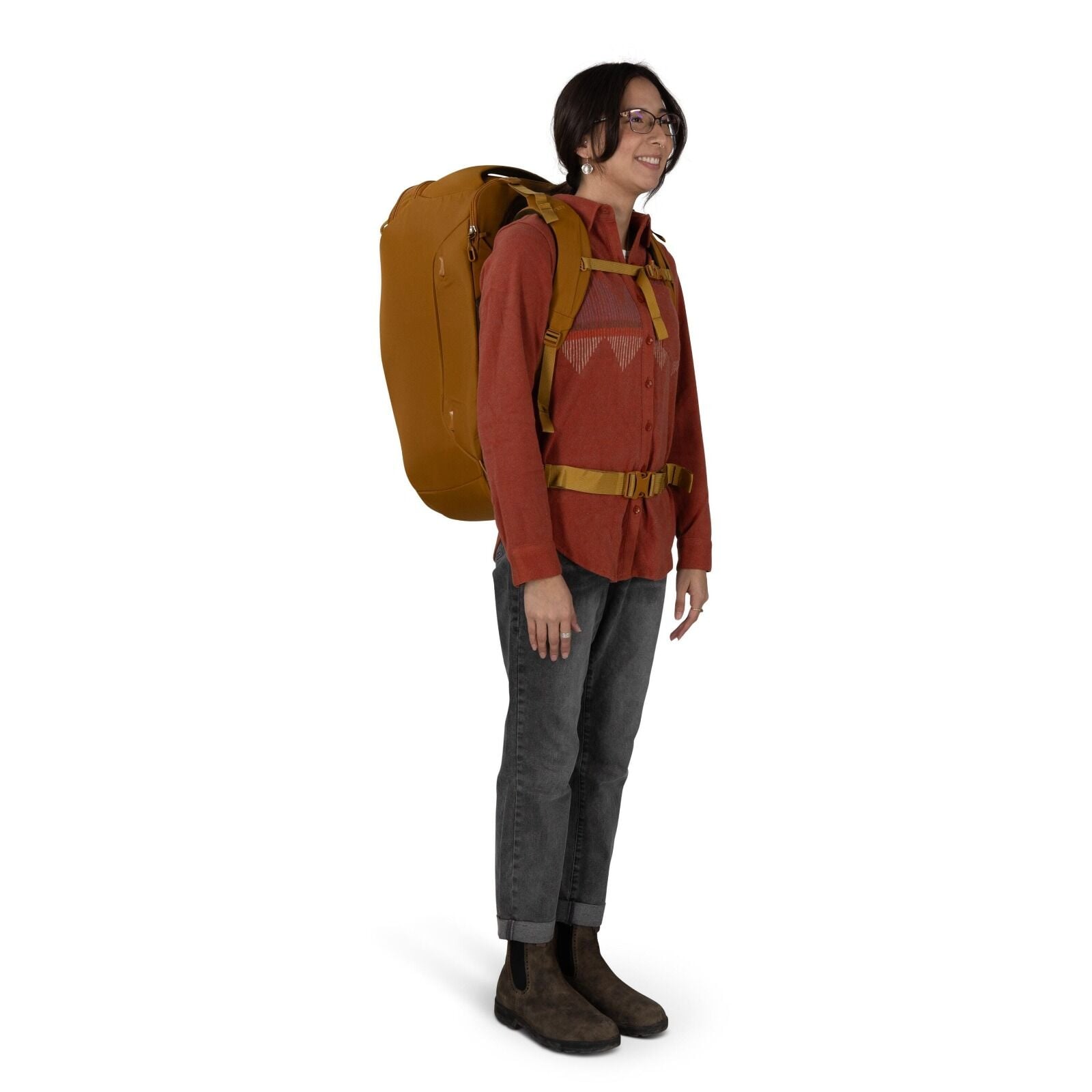 A person is standing in profile wearing a large mustard-colored backpack with padded shoulder straps and a sternum and hip belt. They are dressed in a rust-red button-up shirt with a subtle geometric graphic, cuffed dark gray jeans, and brown Chelsea-styl