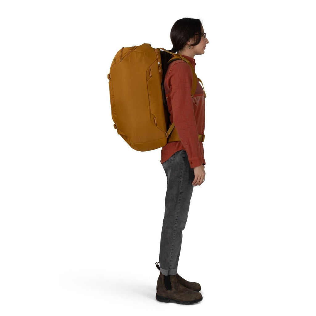 A person is standing in profile wearing a large mustard-yellow travel backpack. They are dressed in a rust-colored long-sleeve shirt, dark gray jeans with a cuff, and brown Chelsea-style boots. The backpack has multiple grab handles, side compression stra