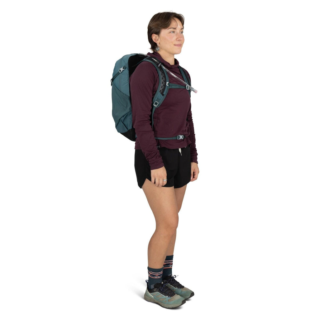 The image shows a person dressed in outdoor hiking attire. They are wearing a burgundy long-sleeve shirt and black shorts, along with blue and pink striped socks. On their feet, they have teal hiking shoes. The person is carrying a teal and black backpack