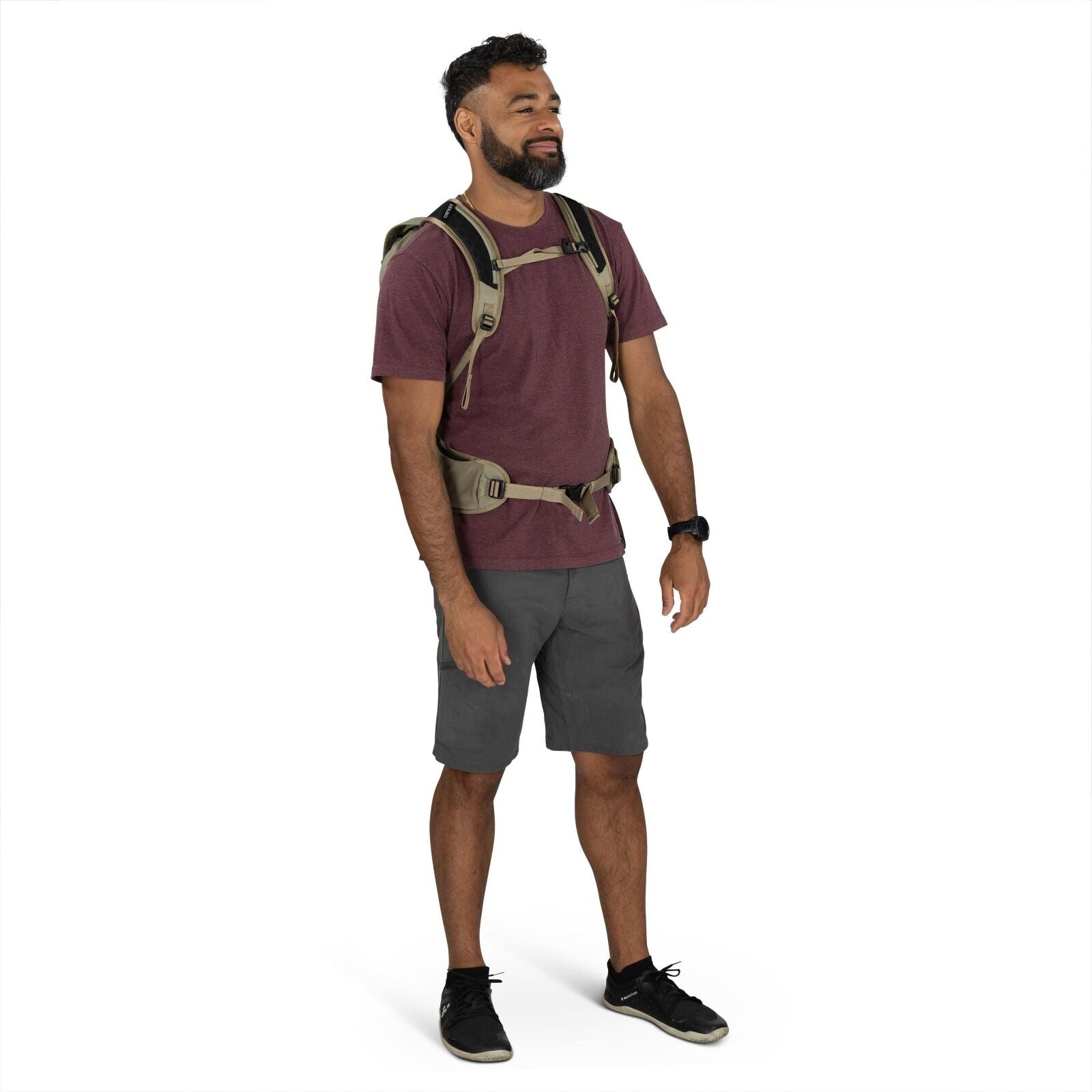 The image shows a man standing against a plain white background. He is wearing a burgundy t-shirt paired with dark gray shorts and black shoes. The man is equipped with an olive-green tactical-style backpack that has multiple straps, including a chest and