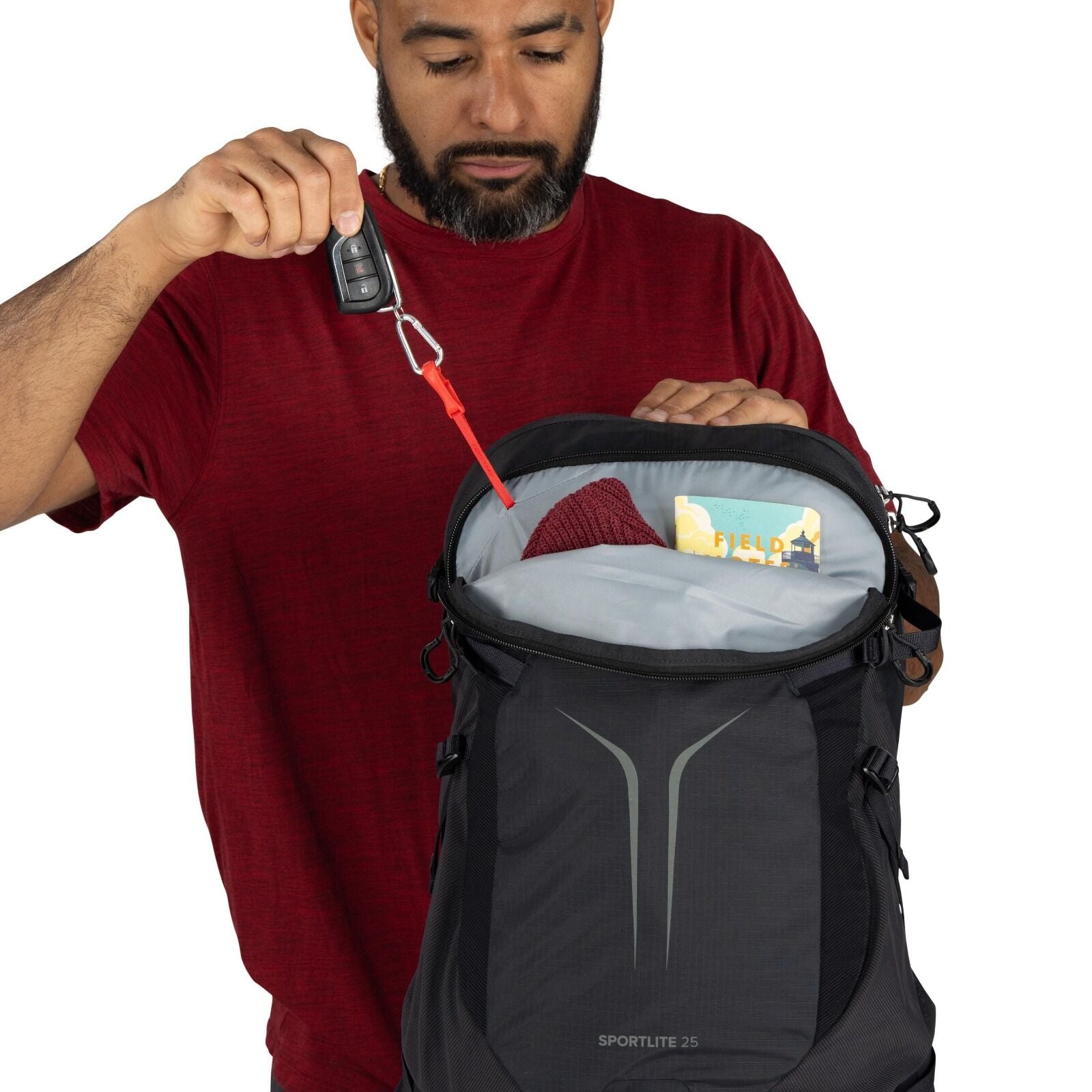 The image features a person wearing a red shirt, holding open a black backpack with "SPORTLITE 25" branding on it. The backpack is partially open, revealing its contents, which include a red knit item, likely a hat or scarf, and a yellow and blue booklet