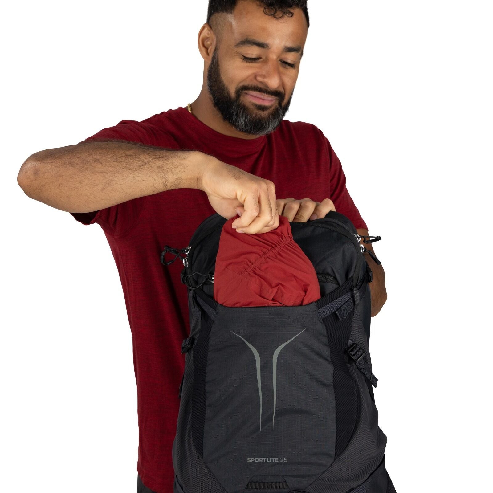 The image shows a person wearing a red short-sleeved shirt and holding a backpack. The backpack is black with a front pocket, prominently displaying the logo "SPORTLITE 25" near the bottom. The person is pulling a red garment out of the backpack. The back