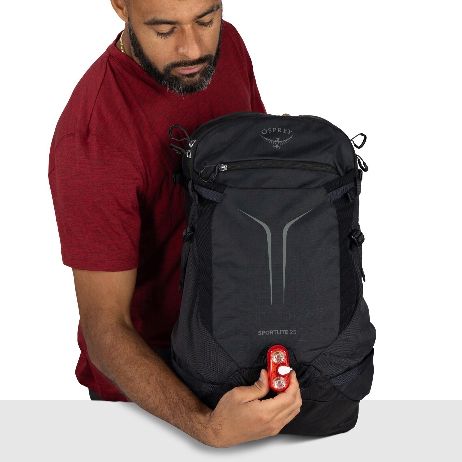 The image shows a person wearing a red shirt holding a black Osprey backpack. The backpack is marked with "SPORTLITE 25" on its front, indicating its model. The person is holding a red safety light, which is attached to the lower part of the backpack. The
