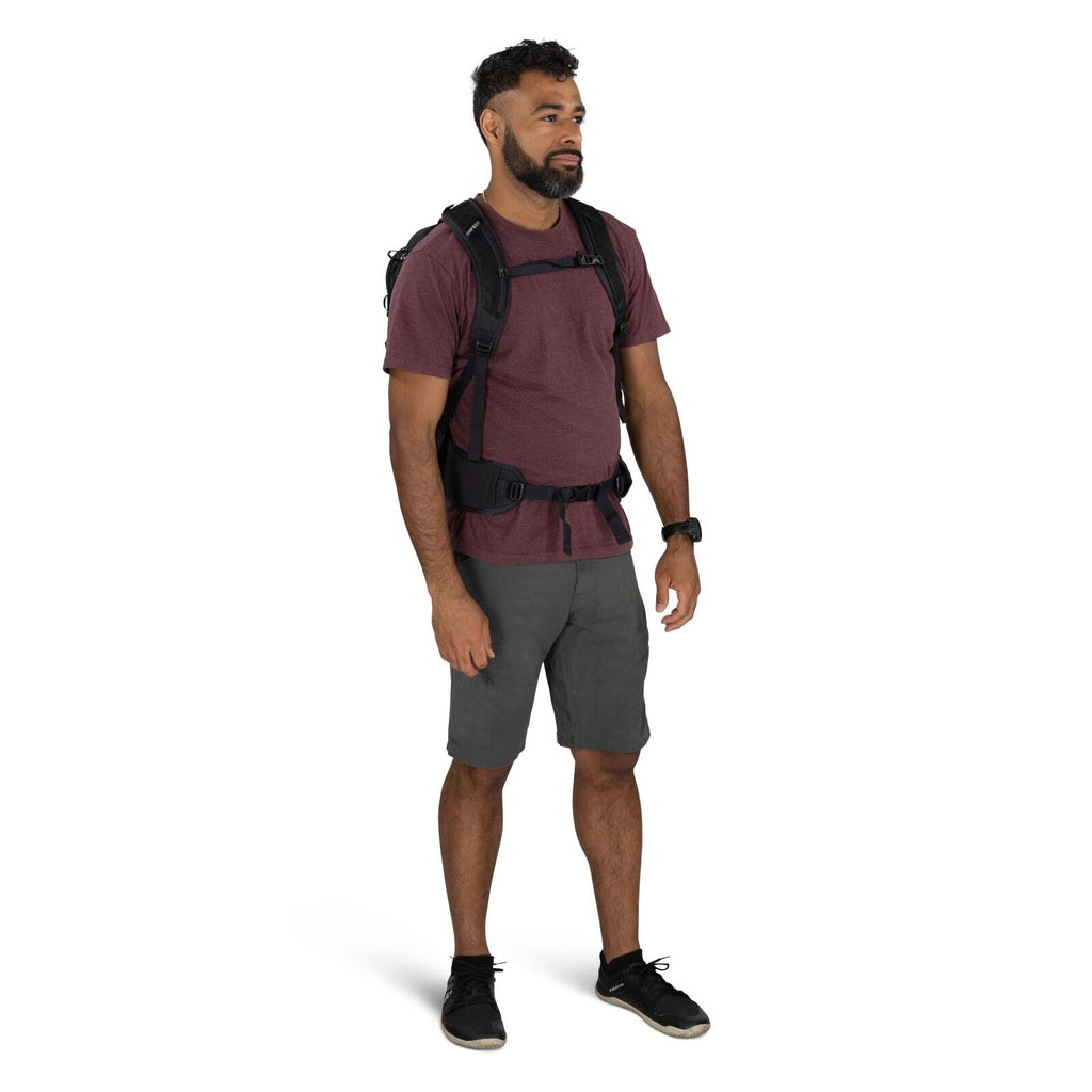 The image shows a man standing and wearing casual outdoor attire, suitable for hiking or adventure activities. He is dressed in a maroon t-shirt paired with gray shorts, and on his feet are black athletic shoes with laces. He is equipped with a black back