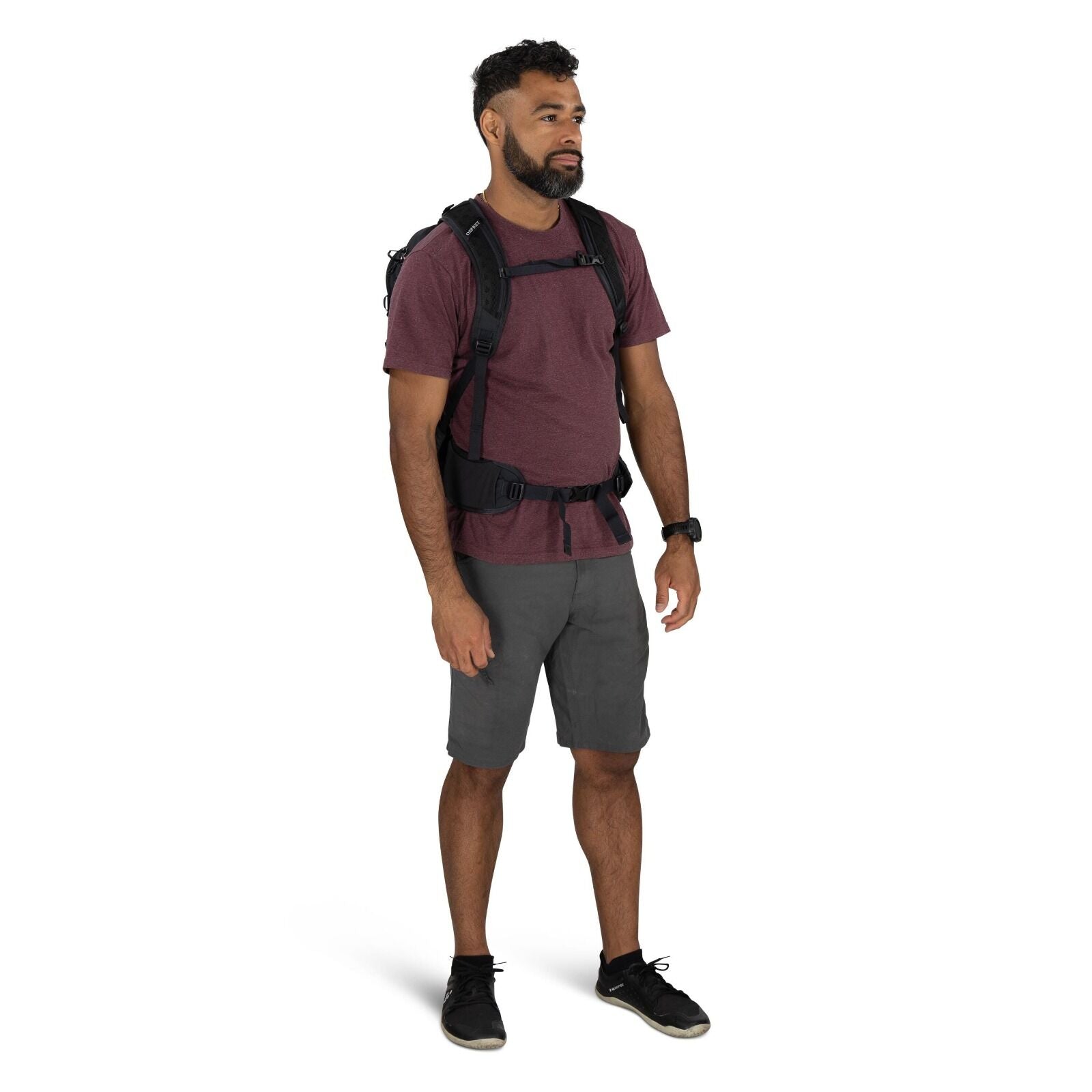 The image shows a man standing and wearing casual outdoor attire, suitable for hiking or adventure activities. He is dressed in a maroon t-shirt paired with gray shorts, and on his feet are black athletic shoes with laces. He is equipped with a black back