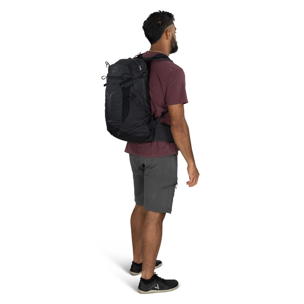 The image depicts a man standing and facing slightly to the side, wearing a casual outfit suitable for outdoor activities. He is dressed in a burgundy short-sleeve t-shirt, grey cargo shorts, and black athletic shoes. The man has a black Osprey backpack o