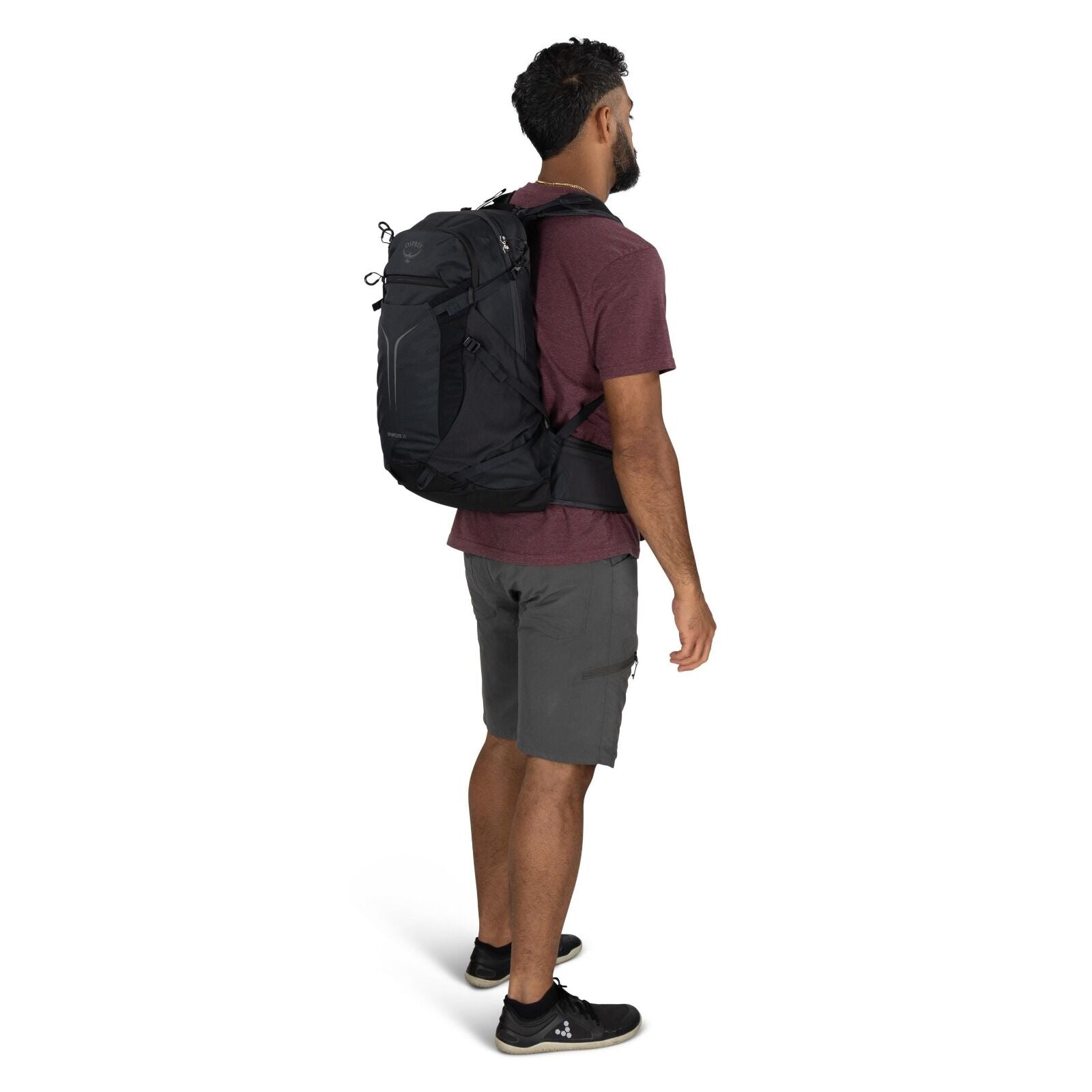 The image depicts a man standing and facing slightly to the side, wearing a casual outfit suitable for outdoor activities. He is dressed in a burgundy short-sleeve t-shirt, grey cargo shorts, and black athletic shoes. The man has a black Osprey backpack o