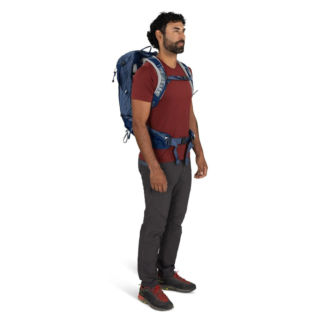 The image shows a person standing in a relaxed pose, wearing hiking or outdoor attire. The individual is dressed in a maroon short-sleeved t-shirt, charcoal gray pants, and black and red hiking shoes. They are equipped with a blue backpack that has variou