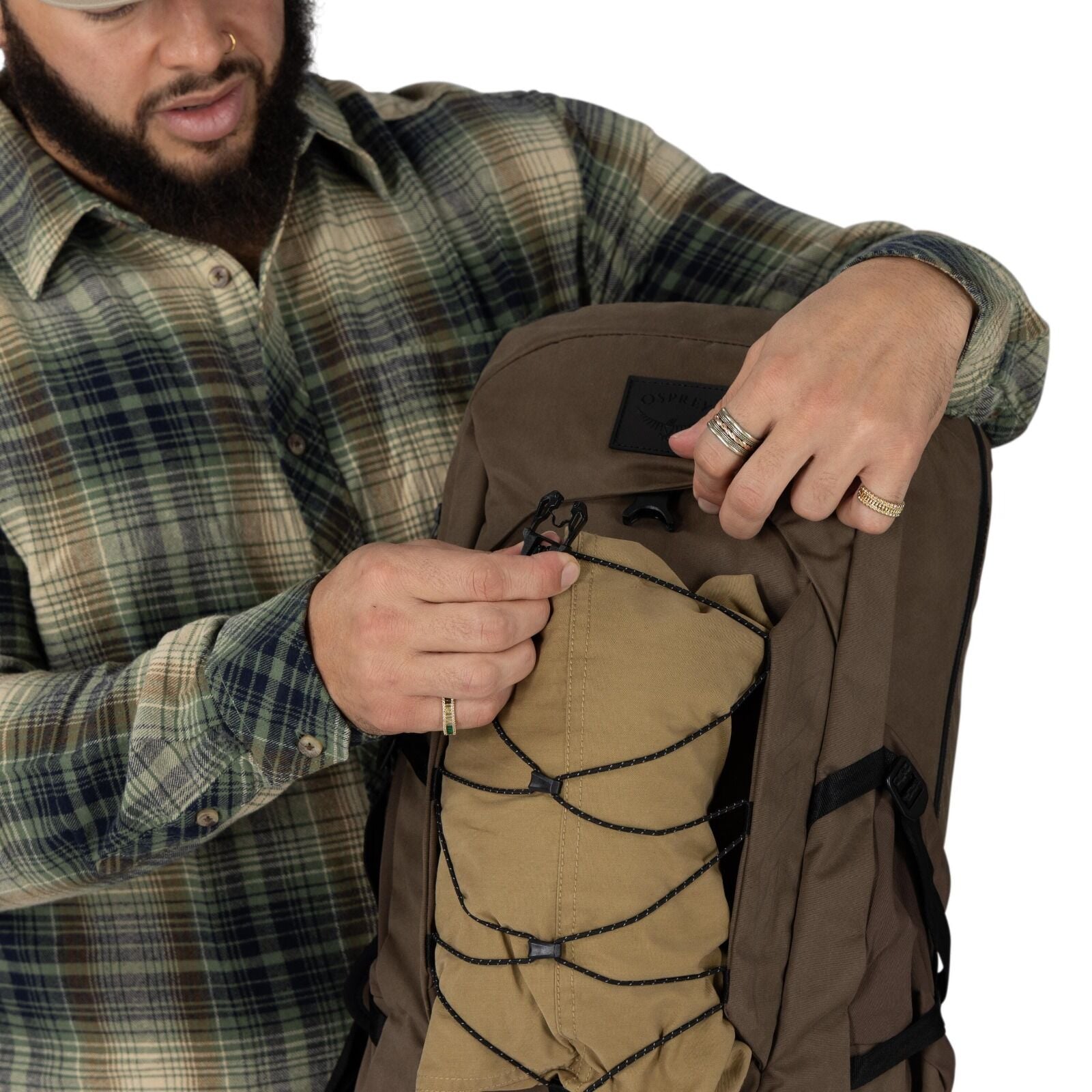 A person wearing a green and brown plaid shirt adjusts the front cord system on a brown Osprey backpack. The pack has a tan shove-it pocket secured with a black bungee lattice and cord locks. The individual's face is cropped out, but their hands with seve