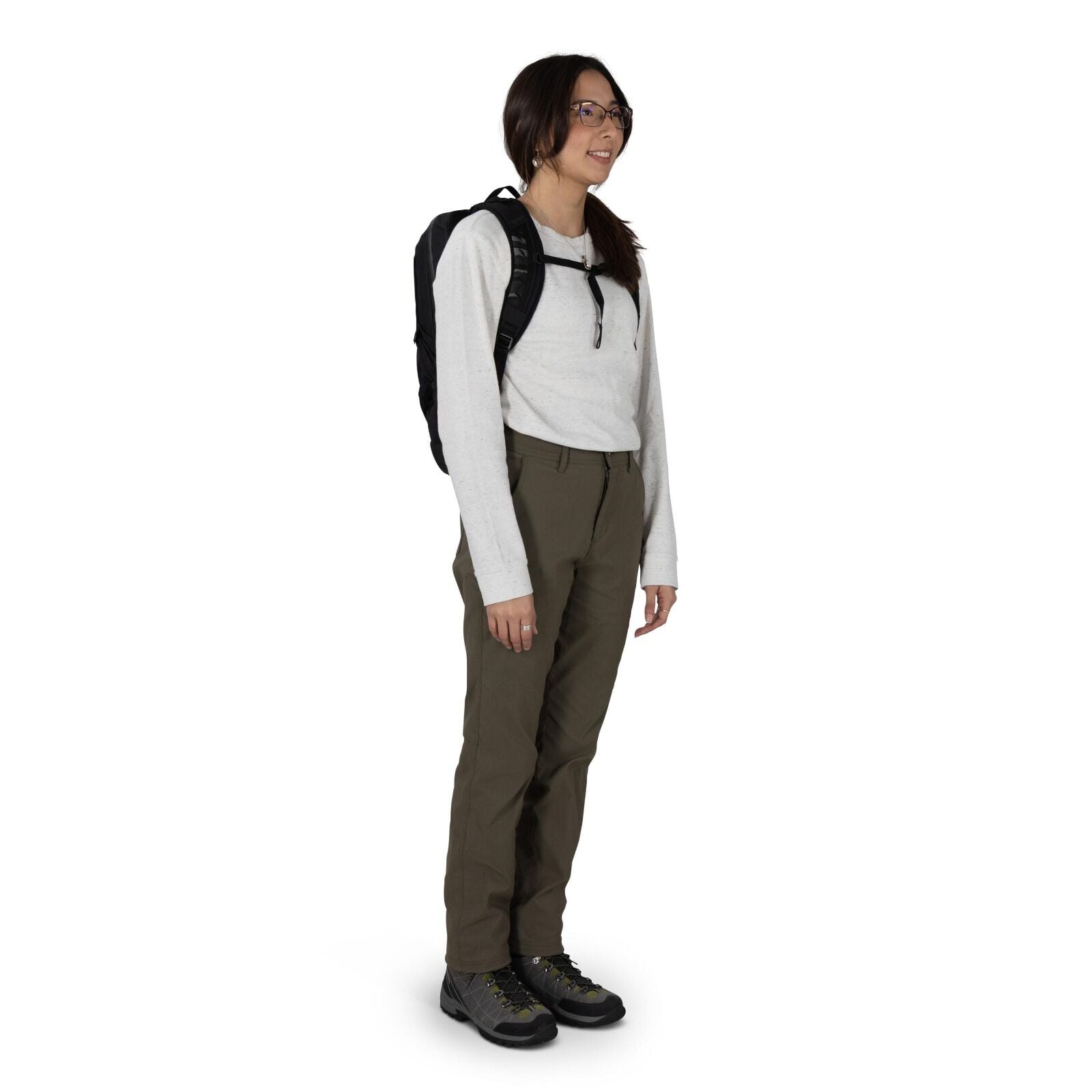 A person stands in a studio setting wearing outdoor clothing and a small black backpack. They have on a light gray long_sleeve shirt tucked into olive green hiking pants. The outfit is completed with gray hiking shoes featuring black toe caps and lime acc