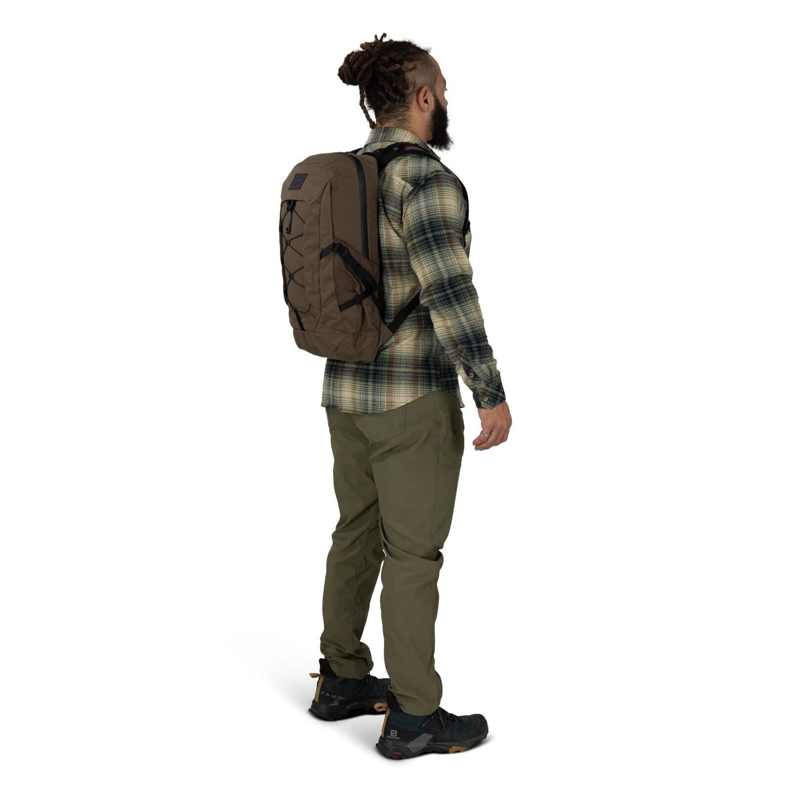 A person stands in a three-quarter side pose wearing outdoor clothing and a backpack. They have on a green plaid flannel shirt, olive cargo-style pants, and black trail shoes with rugged soles. The backpack is brown with black straps and a front bungee co