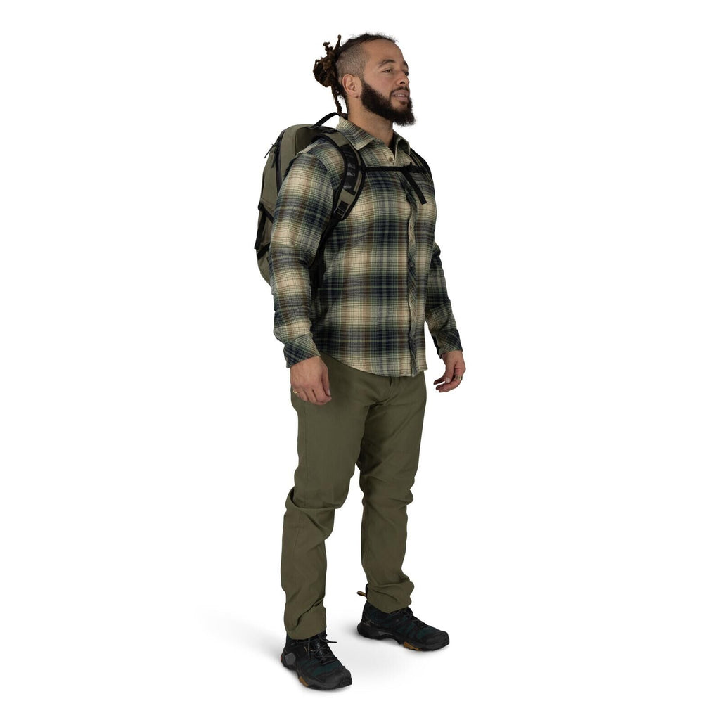 A person stands against a plain white background wearing outdoor clothing and a backpack. They have on a green and beige plaid button-up shirt, olive hiking pants, and black trail shoes. The backpack is brown with black straps, and a chest strap is clippe