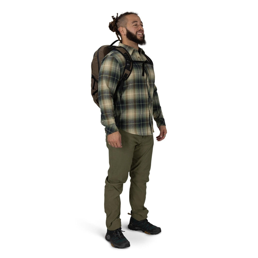 A person stands against a plain white background wearing outdoor clothing and a backpack. They have on a green and beige plaid button-up shirt, olive hiking pants, and black trail shoes. The backpack is brown with black straps, and a chest strap is clippe