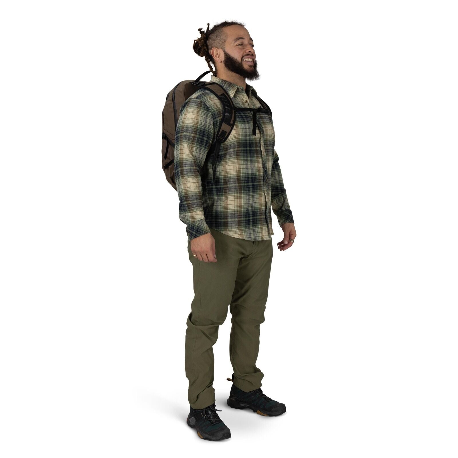 A person stands against a plain white background wearing outdoor clothing and a backpack. They have on a green and beige plaid button-up shirt, olive hiking pants, and black trail shoes. The backpack is brown with black straps, and a chest strap is clippe