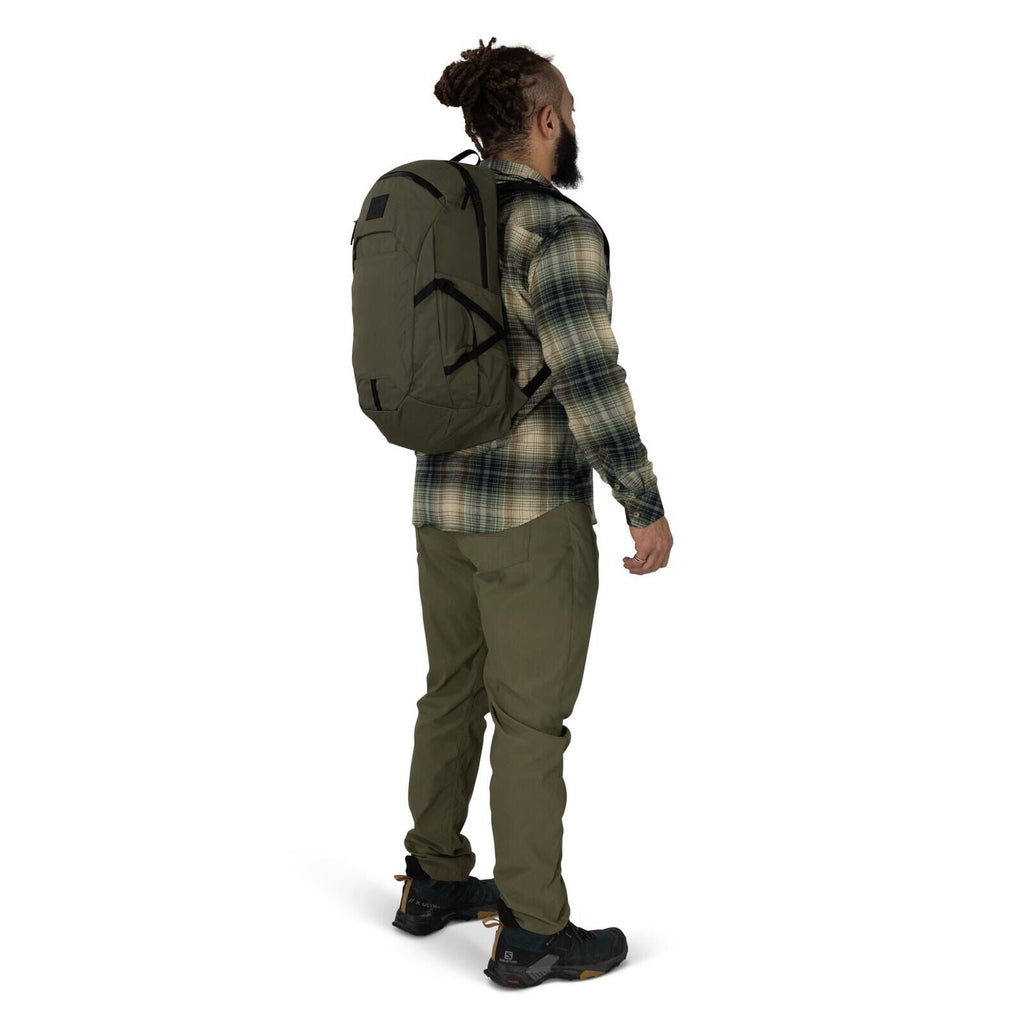A person stands in a three-quarter rear view wearing outdoor clothing and a large brown backpack. They have on a green plaid flannel shirt, olive cargo-style pants, and black trail shoes with rugged soles. Their hair is tied up in a bun, and they have a b