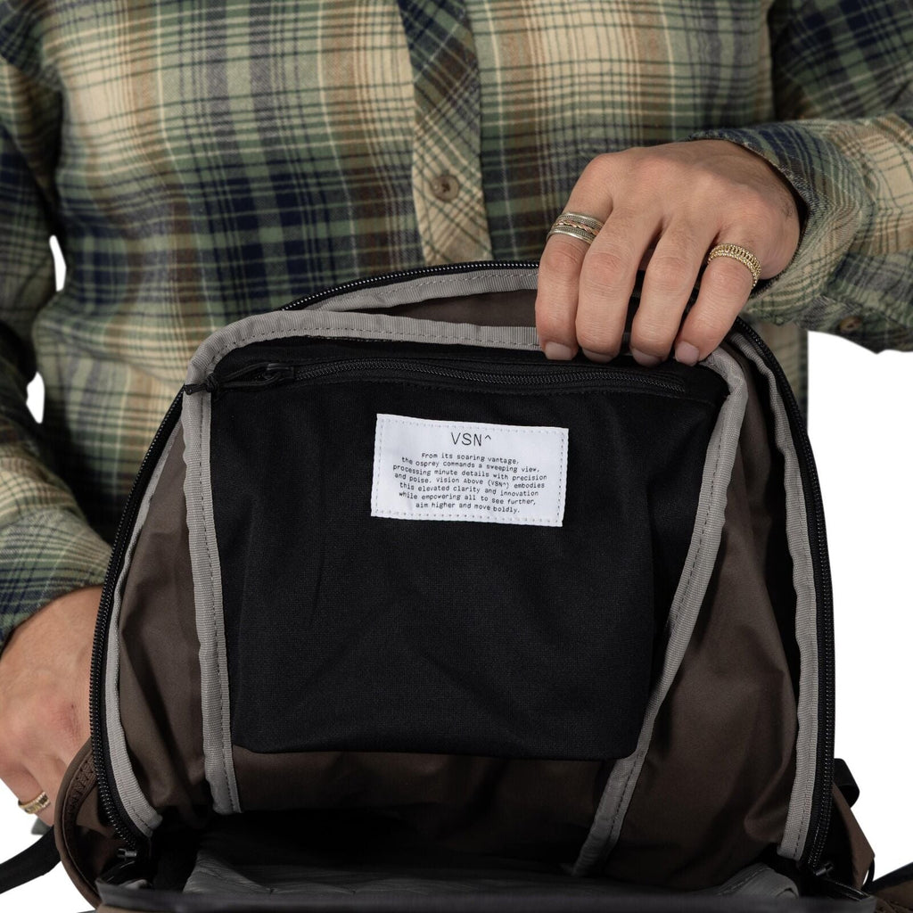 A person in a green plaid shirt holds open a brown backpack to show the inside front panel which has a description about the VSN, Vision Above, product that leads in innovation and technological advancement.