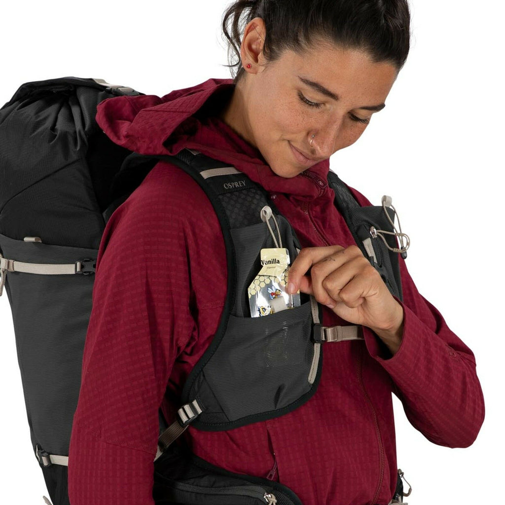 A person in a red jacket and backpack smiles while placing a vanilla snack into a black front pocket. The tone is adventurous and content.