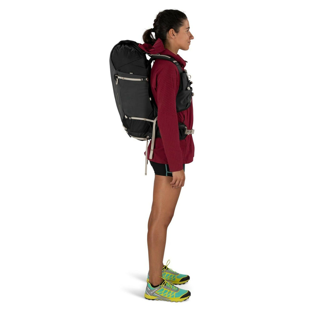 Profile of a woman standing wearing a large black backpack and a red jacket. She has on athletic clothing and green trail running shoes.