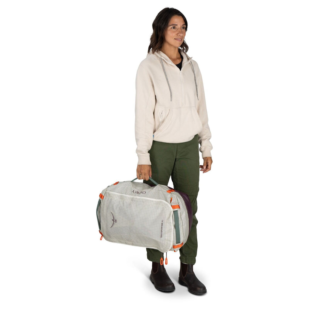 The image features a person standing and holding a duffel bag in one hand. The individual is dressed in a light-colored hooded sweatshirt, olive green pants, and brown boots. The duffel bag is predominantly light-colored with purple ends and features oran