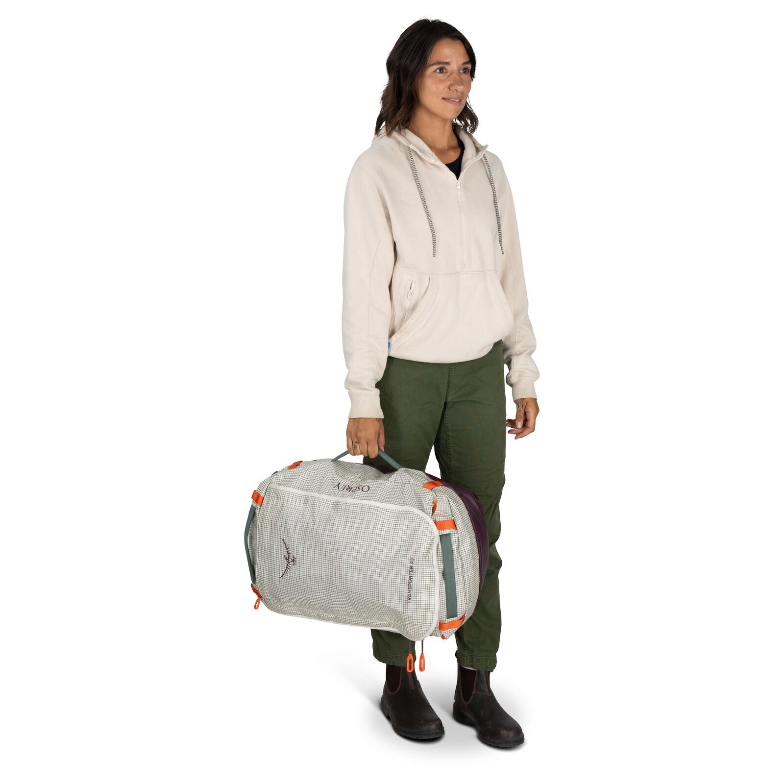 The image features a person standing and holding a duffel bag in one hand. The individual is dressed in a light-colored hooded sweatshirt, olive green pants, and brown boots. The duffel bag is predominantly light-colored with purple ends and features oran