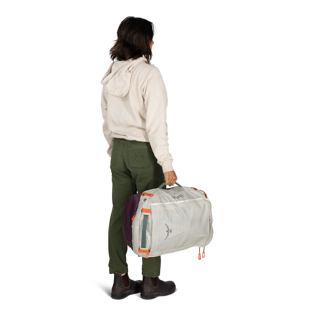 The image features a person standing and holding a duffel bag in one hand. The individual is dressed in a light-colored hooded sweatshirt, olive green pants, and brown boots. The duffel bag is predominantly light-colored with purple ends and features oran