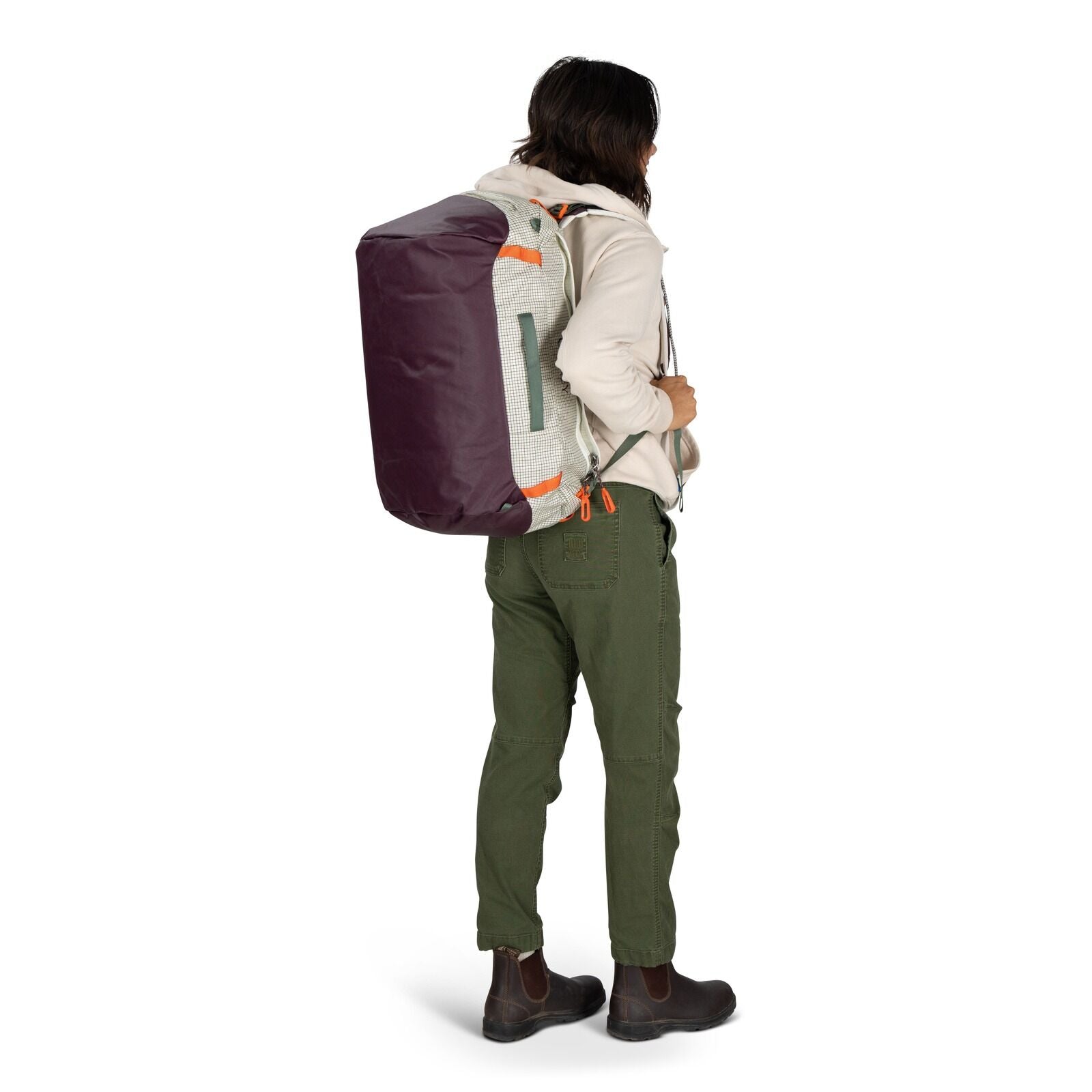 The image features a person standing and holding a duffel bag in one hand. The individual is dressed in a light-colored hooded sweatshirt, olive green pants, and brown boots. The duffel bag is predominantly light-colored with purple ends and features oran