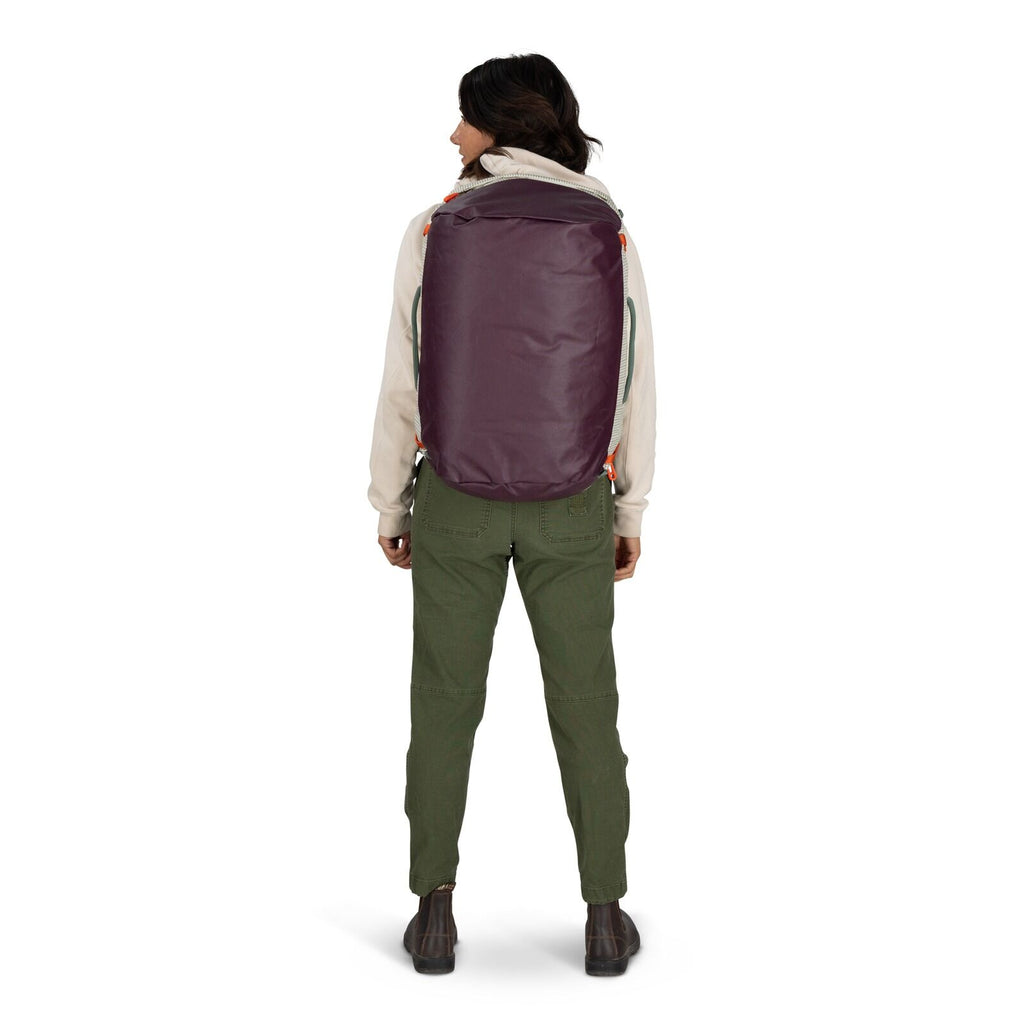The image features a person standing and holding a duffel bag in one hand. The individual is dressed in a light-colored hooded sweatshirt, olive green pants, and brown boots. The duffel bag is predominantly light-colored with purple ends and features oran