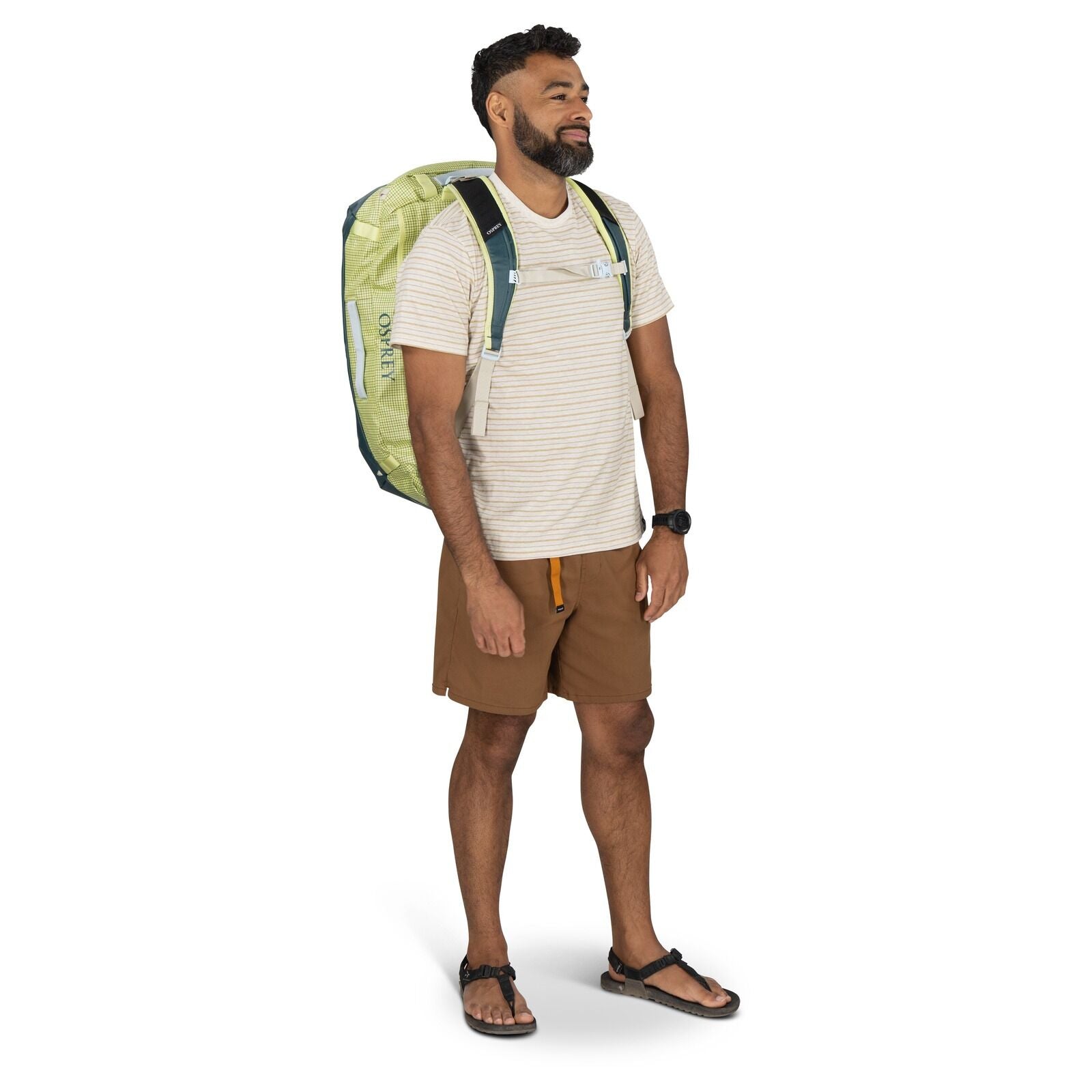 The image shows a man standing upright wearing a light-colored, short-sleeve, striped T-shirt paired with brown shorts. He has a large Osprey brand backpack on his back, which is predominantly light green with dark green accents. On his wrist, he wears a