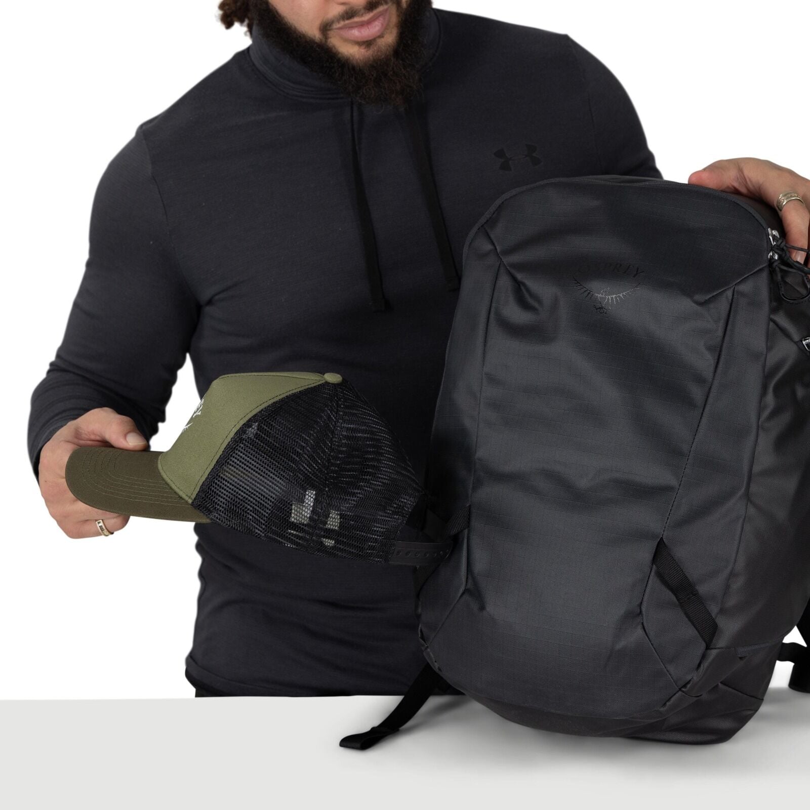 A person wearing a dark hoodie and a gray beanie is opening a black backpack and sliding a laptop into the padded sleeve. The pack's main compartment is unzipped wide, revealing an inner organizer pocket holding a magazine labeled "BOOKS." The backpack ha