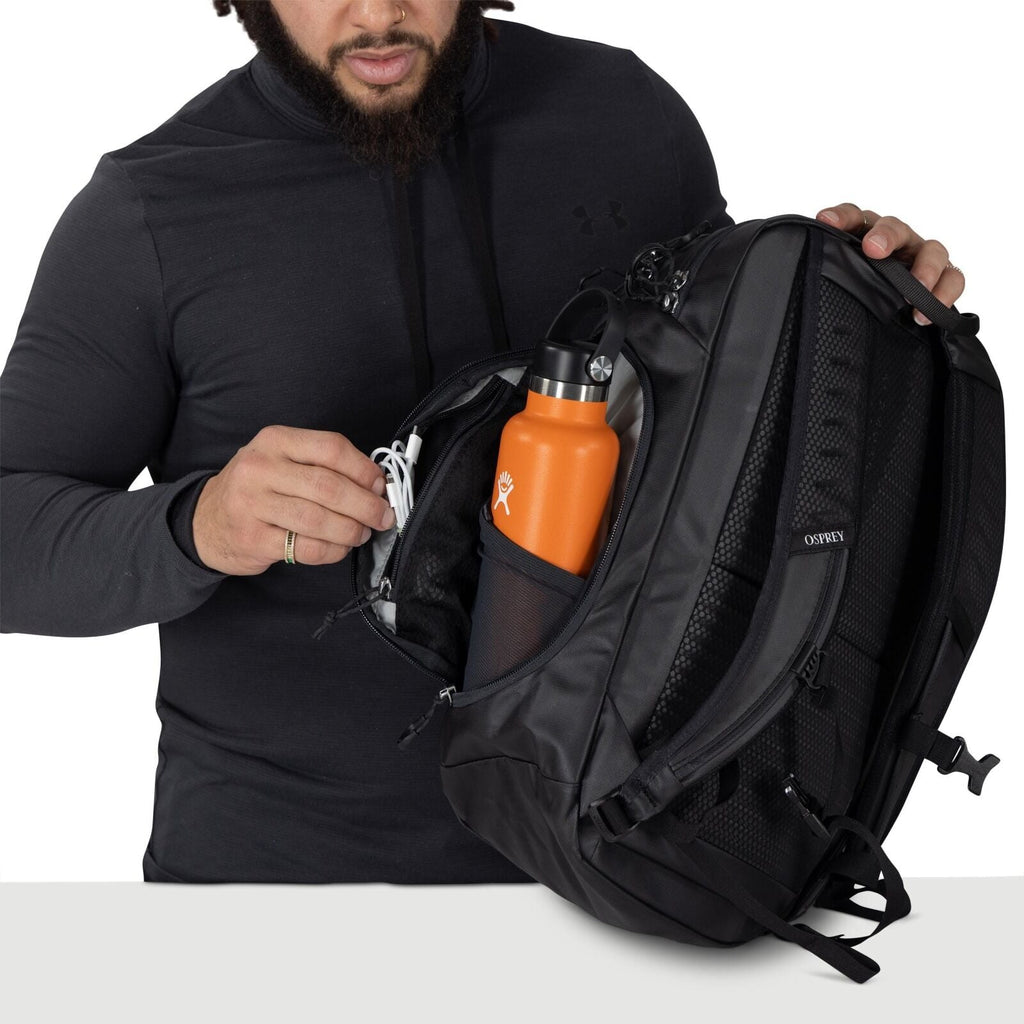 A person wearing a dark hoodie opens a black Osprey backpack. Inside, an orange Hydro Flask bottle sits in a mesh side pocket. The person is organizing a small zippered compartment, holding a white cable or earphones. The bag features padded straps and mu