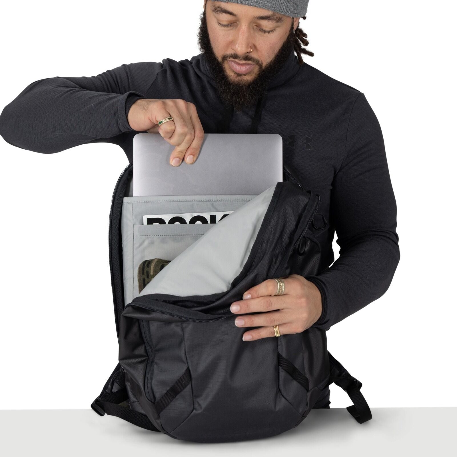 A person wearing a dark Under Armour pullover is holding a sleek black Osprey backpack. With the other hand, they're placing or removing an olive and black mesh trucker hat from the pack. The scene is shot against a plain white background with a table edg