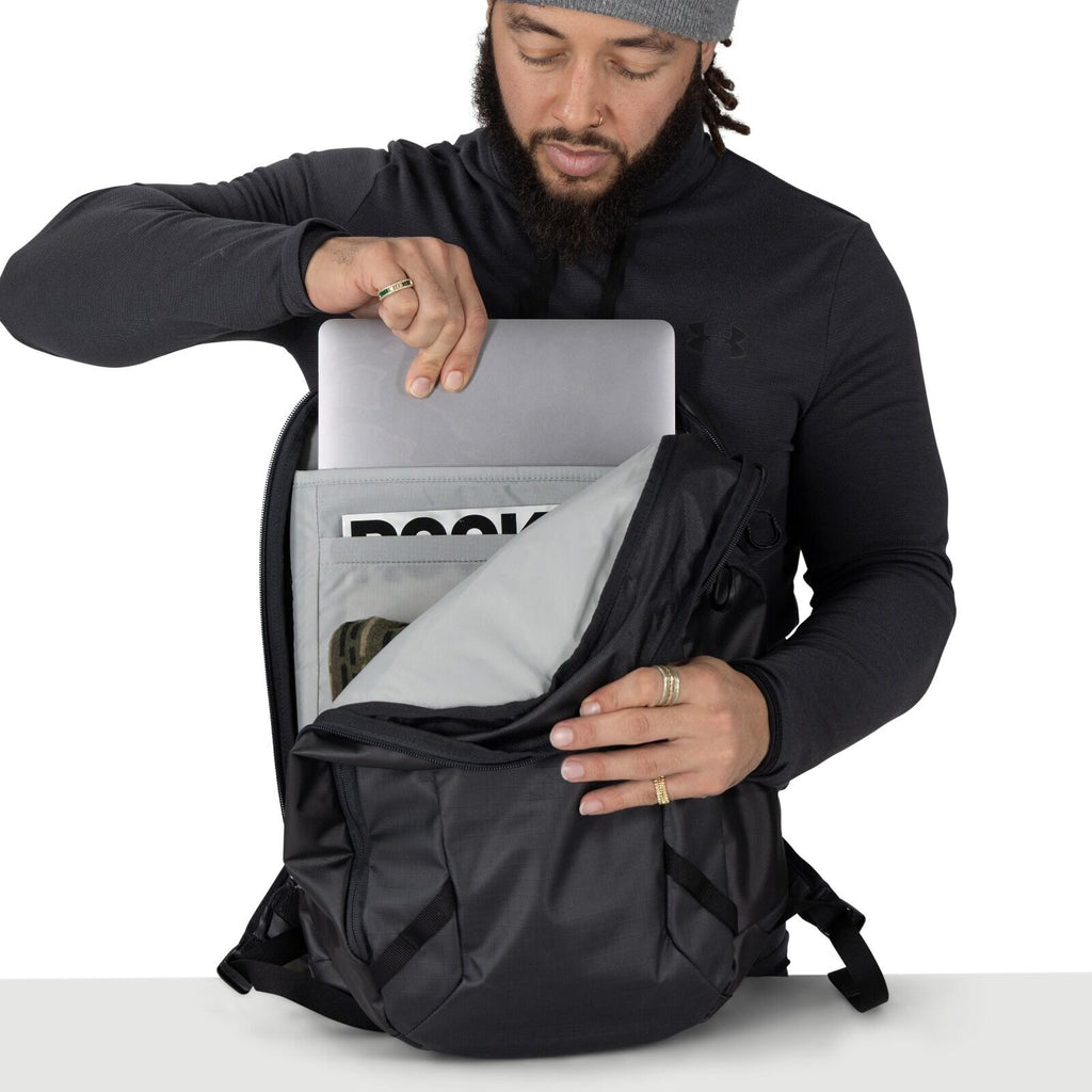 A person wearing a dark hoodie and a gray beanie is opening a black backpack and sliding a laptop into the padded sleeve. The pack's main compartment is unzipped wide, revealing an inner organizer pocket holding a magazine labeled "BOOKS." The backpack ha