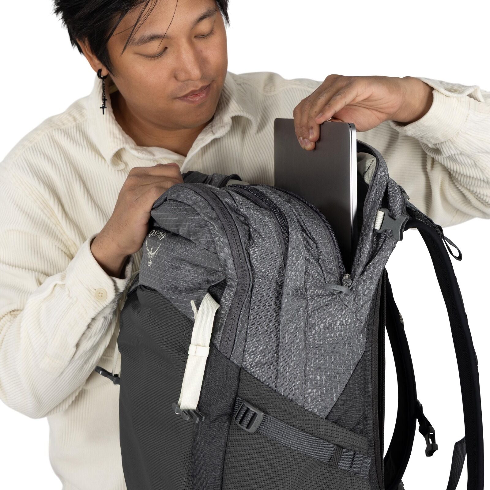 The image shows a person wearing a light-colored button-up shirt and inserting a laptop into a gray Osprey backpack. The backpack features a honeycomb pattern on the fabric and has a large zippered compartment where the laptop is being placed. There is a