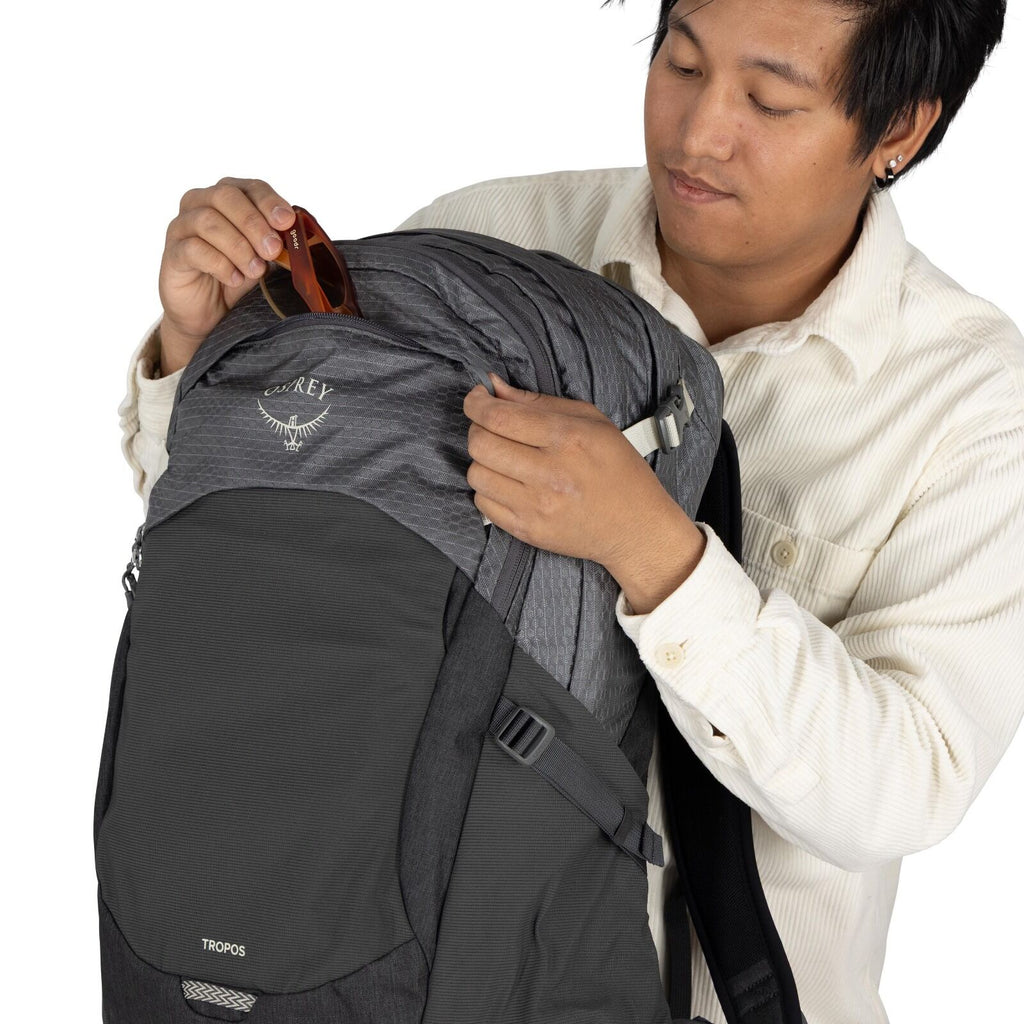 The image shows a person wearing a white, corduroy shirt and holding a grey and black Osprey backpack. The model of the backpack is Tropos, as indicated by the logo and text on the front. The person is placing or removing a pair of brown sunglasses from t
