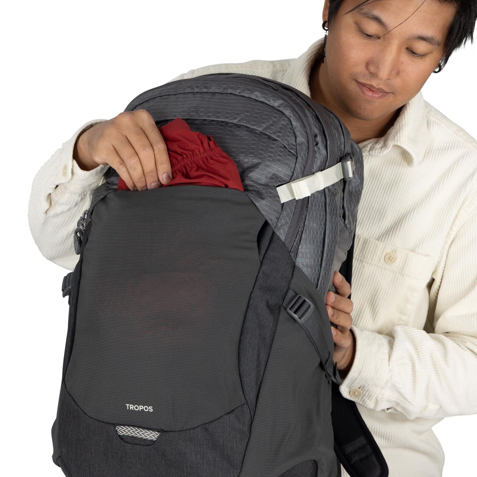 The image shows a person wearing a light-colored, long-sleeved button-up shirt, inserting a red fabric item into the front compartment of a dark gray backpack. The backpack features a design with various compartments, zippers, and adjustable straps. A log