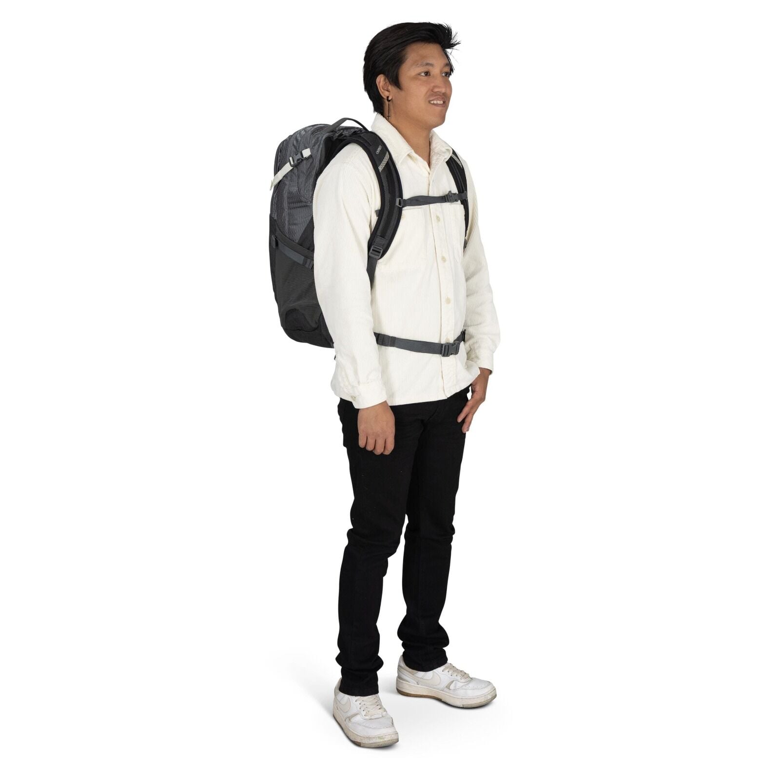 The image shows a person standing and wearing a backpack. The individual is dressed in a casual outfit, featuring a cream-colored shirt, black pants, and white sneakers. The backpack is gray with black accents and includes adjustable straps and buckles fo