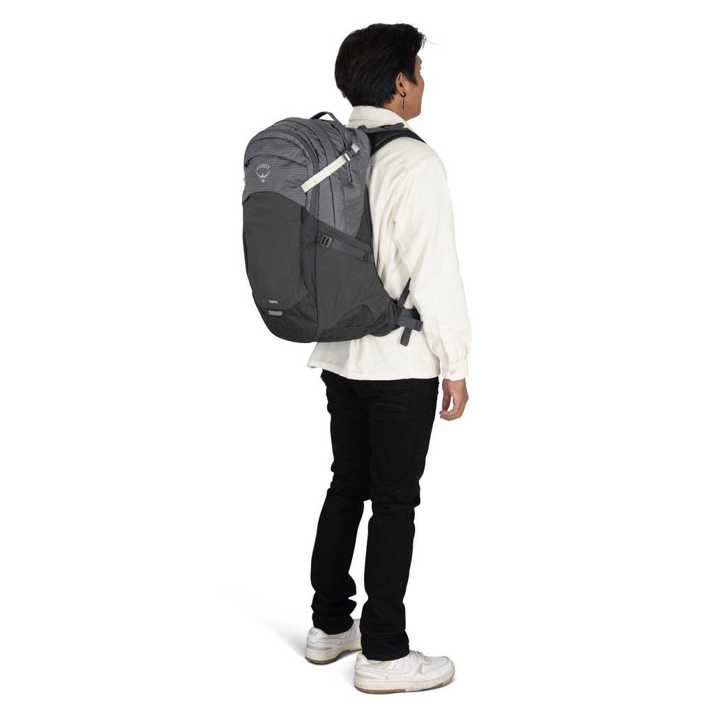 The image shows a person standing, turned slightly to the side, wearing a large, dark gray Osprey backpack. The backpack is designed with multiple compartments and a light gray pattern on its upper section. The person is dressed in a cream-colored shirt,