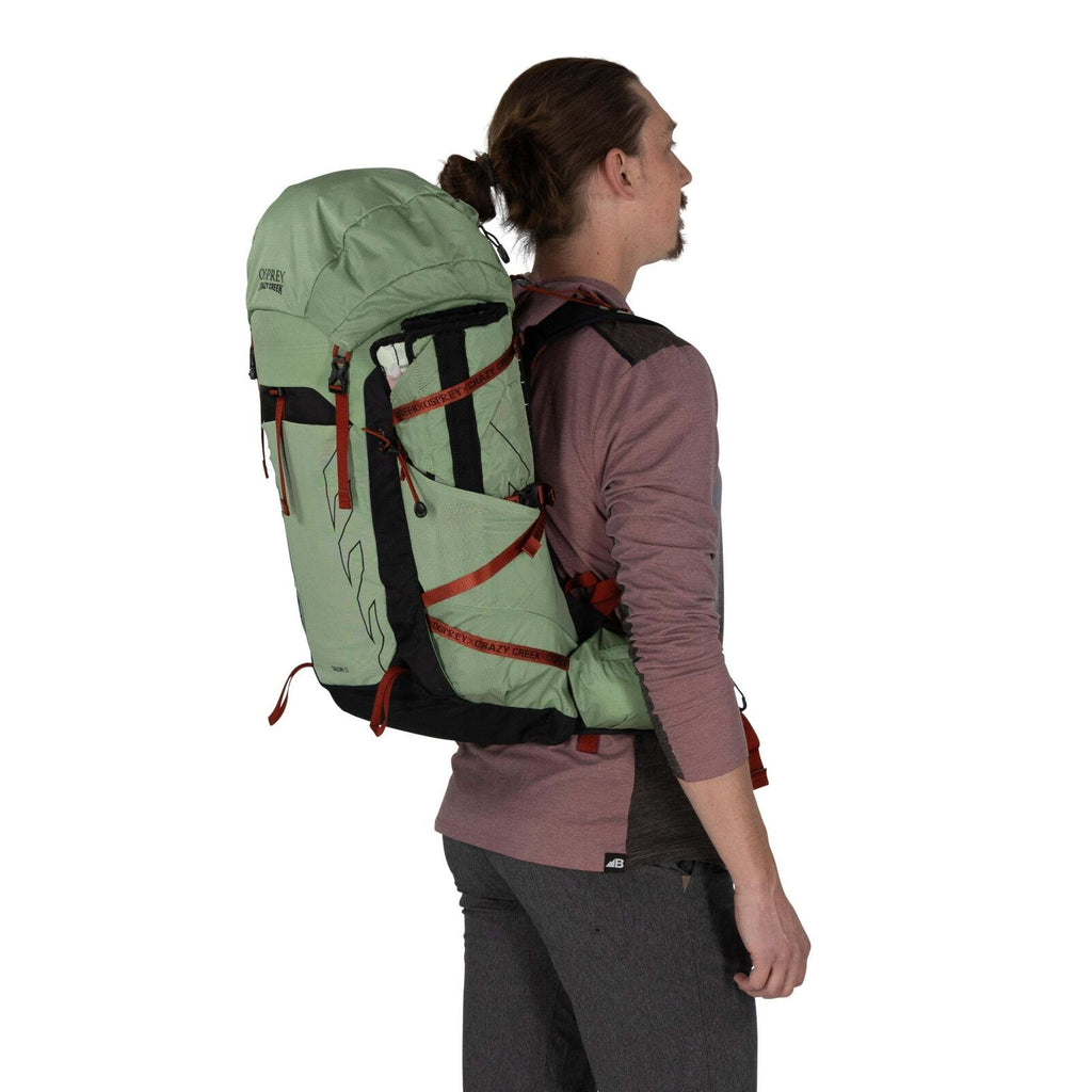 A person wearing a green and black backpack stands sideways, showcasing the pack's design and features against a white background.